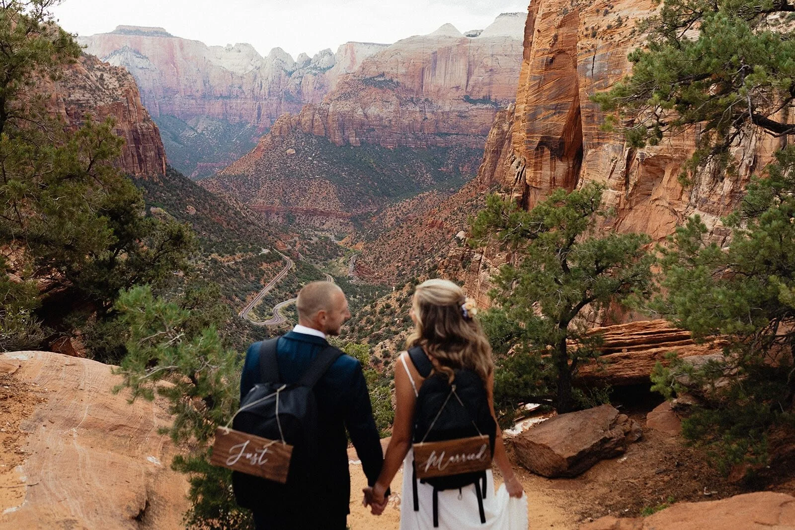 a bride and groom holding hands with backpacks on overlook Zion cliffsides.