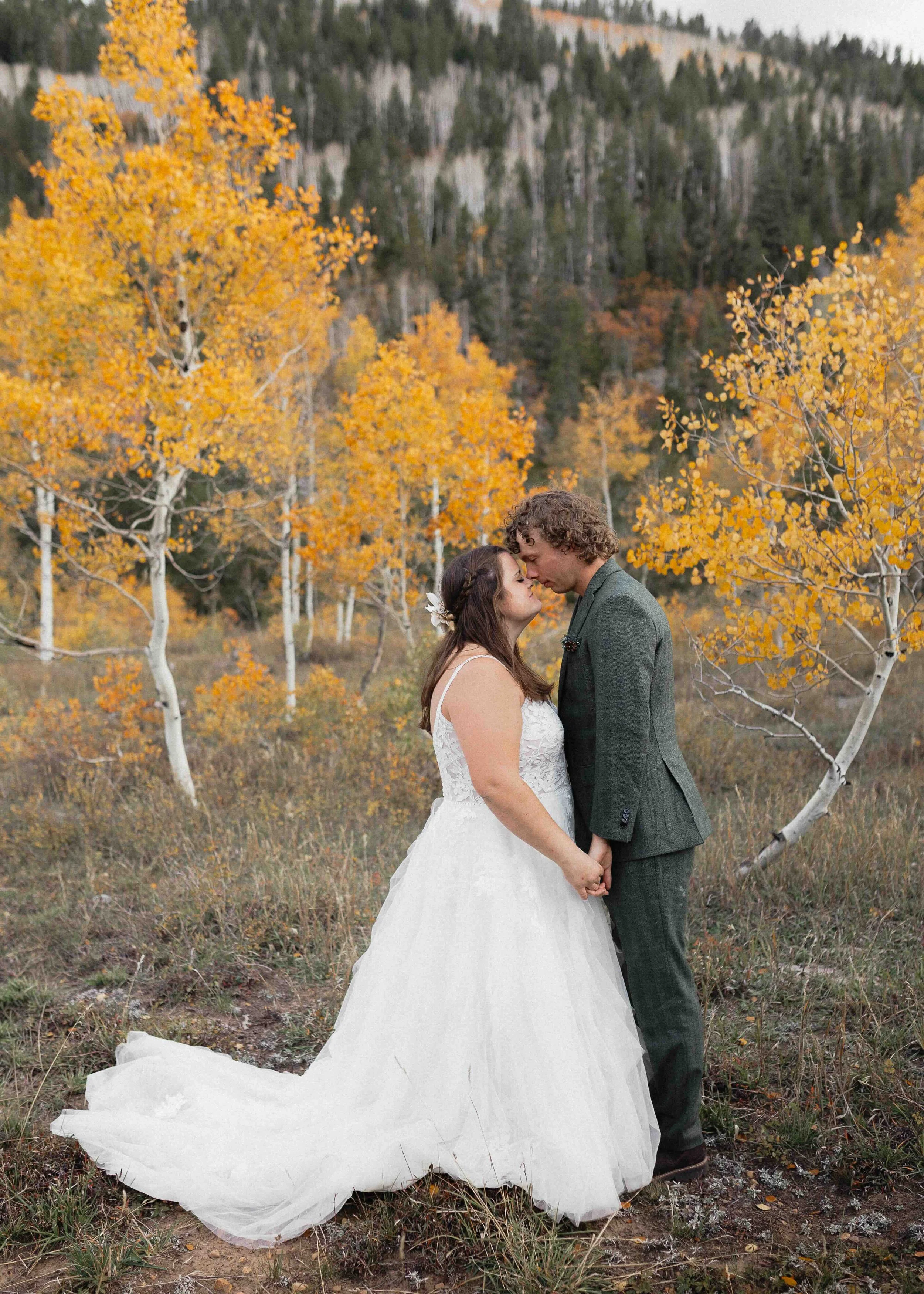 Bride and groom forehead to forehead and holding hands in front of aspen trees with yellow leaves and a mountain.