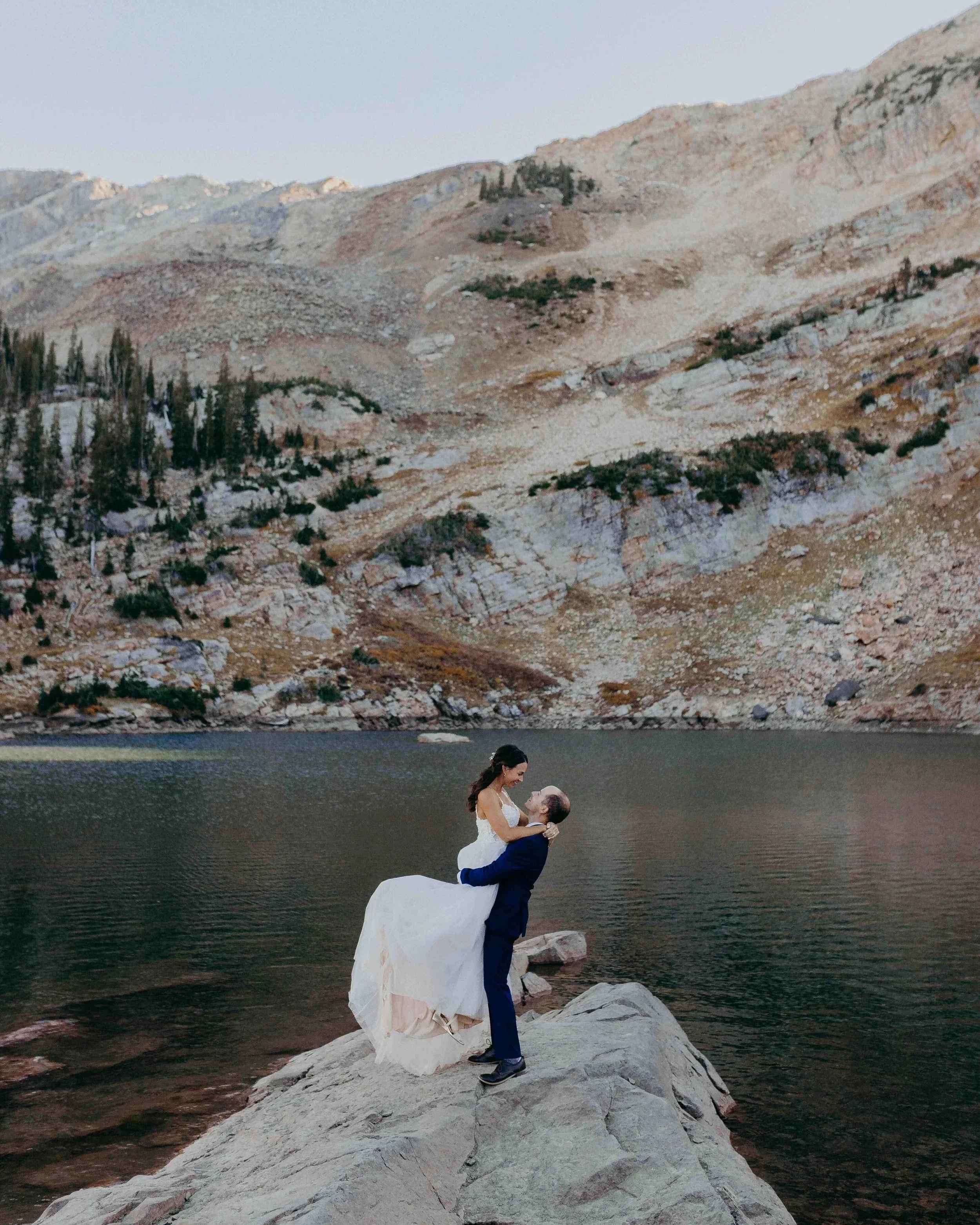 bride and groom on a boulder kissing in front of an alpine lake with rocky mountain in the background.