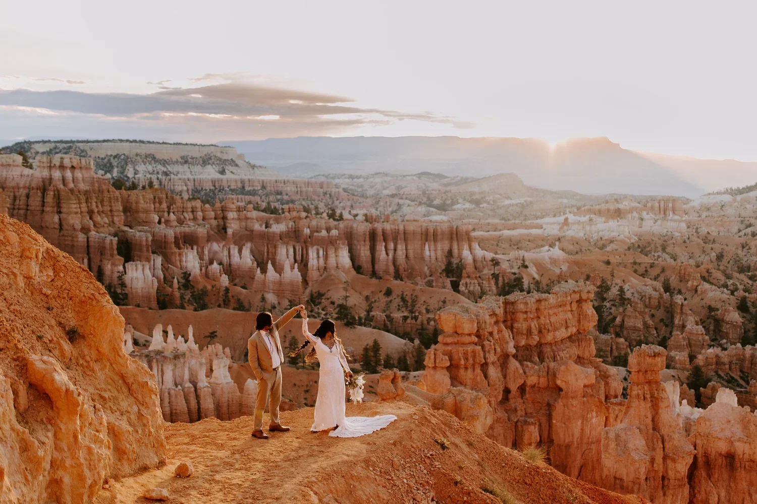 A bride and groom stand on a rocky cliff overlooking the colorful canyon at sunset, with the groom holding the bride's hand.