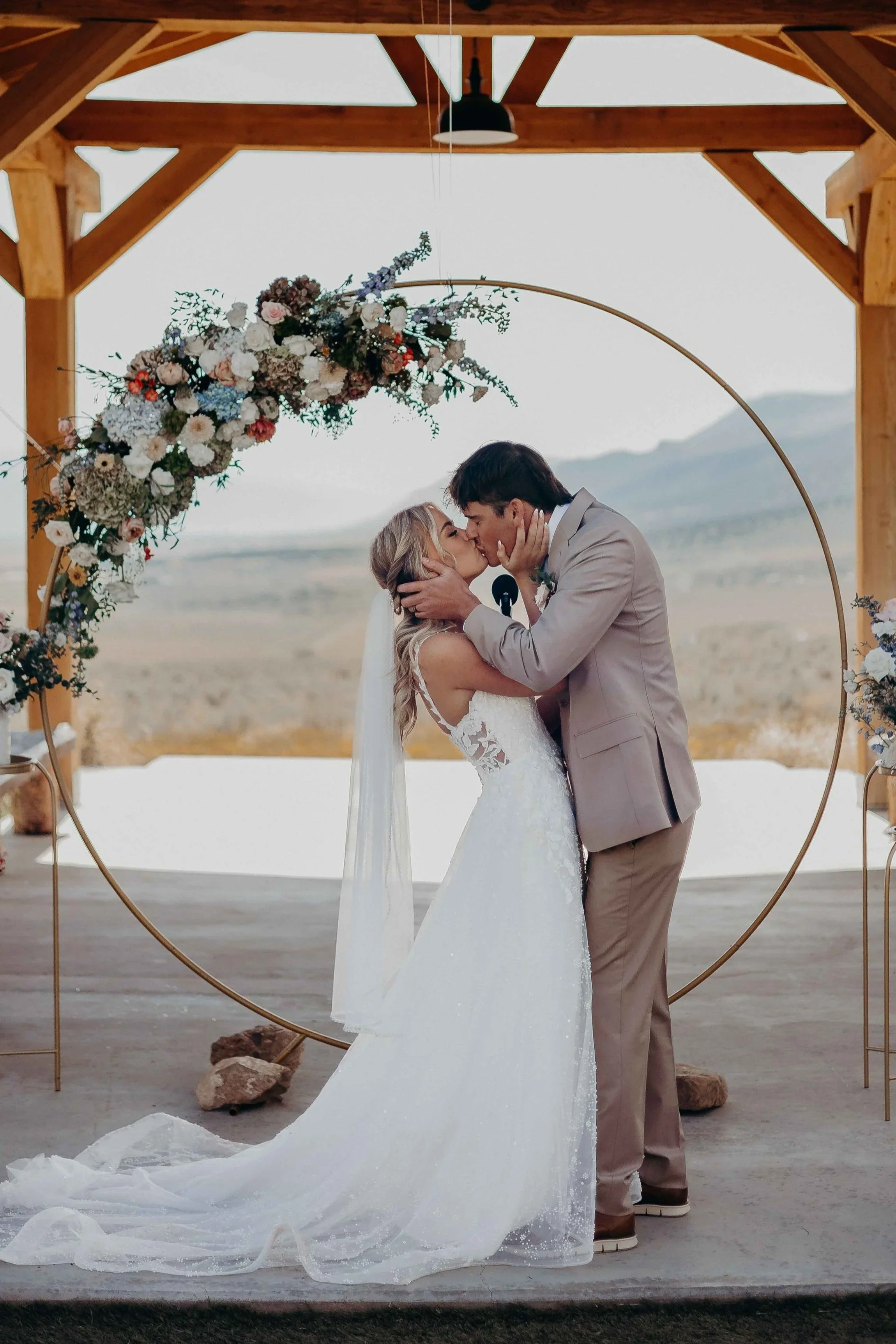 A bride and groom kissing during their outdoor wedding ceremony, with a wooden arch decorated with flowers, set against a landscape background.