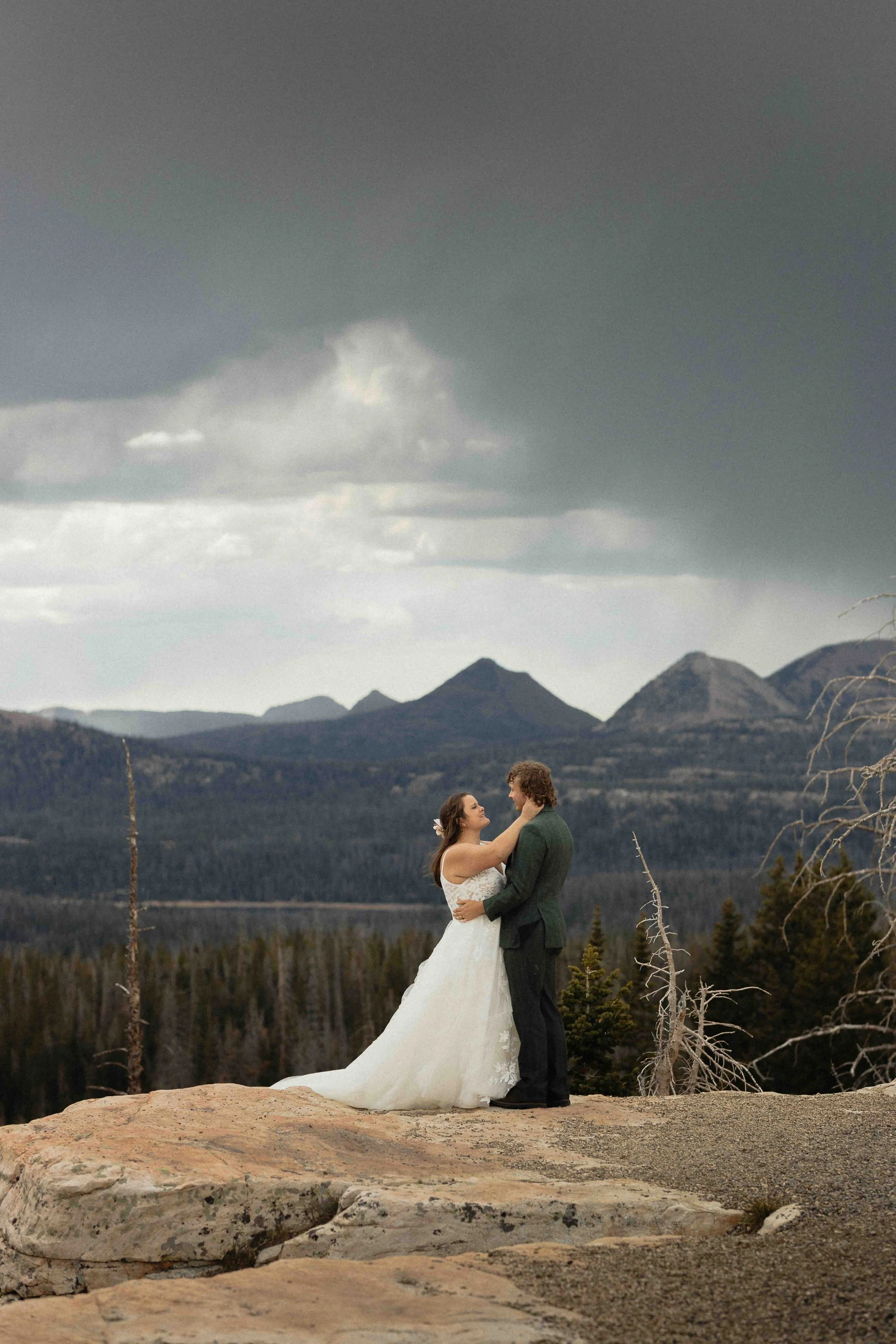 Bride and groom standing on a cliffside with mountains in the background with storm clouds above them.