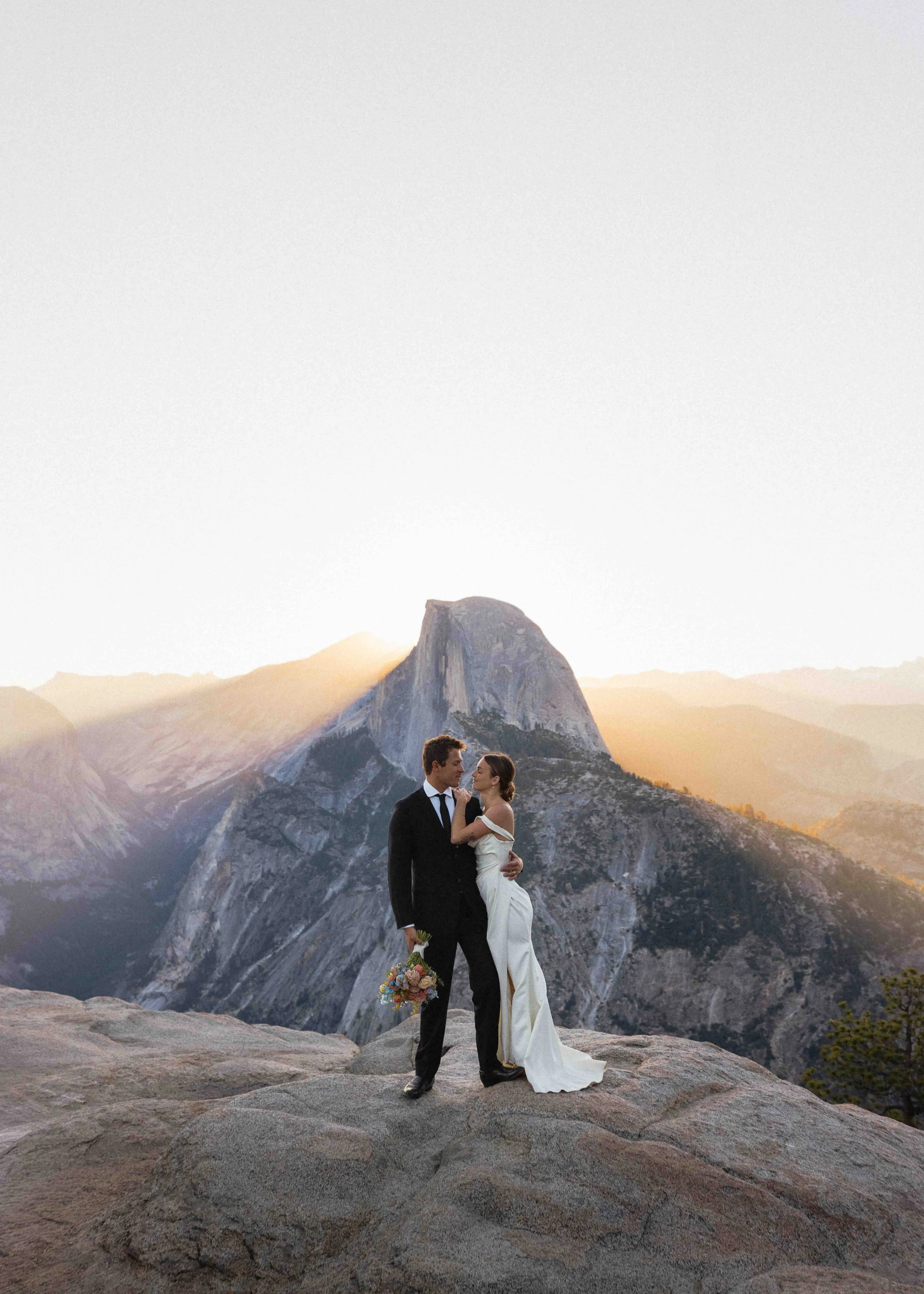 Bride and groom standing at Glacier point in Yosemite national park overlooking the half dome at sunset. Light pours over jutting grey cliff sides in the background.