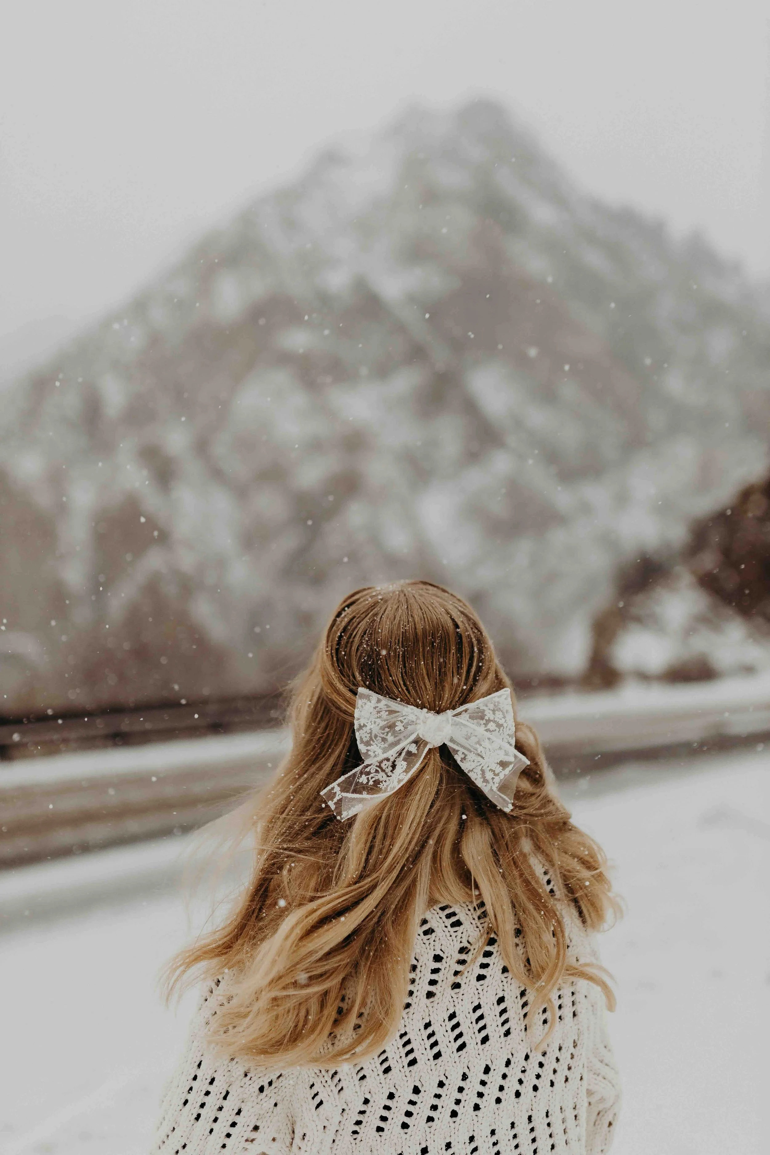 Girl looking out at snowy mountain top with white bow in her hair and a white sweater on.