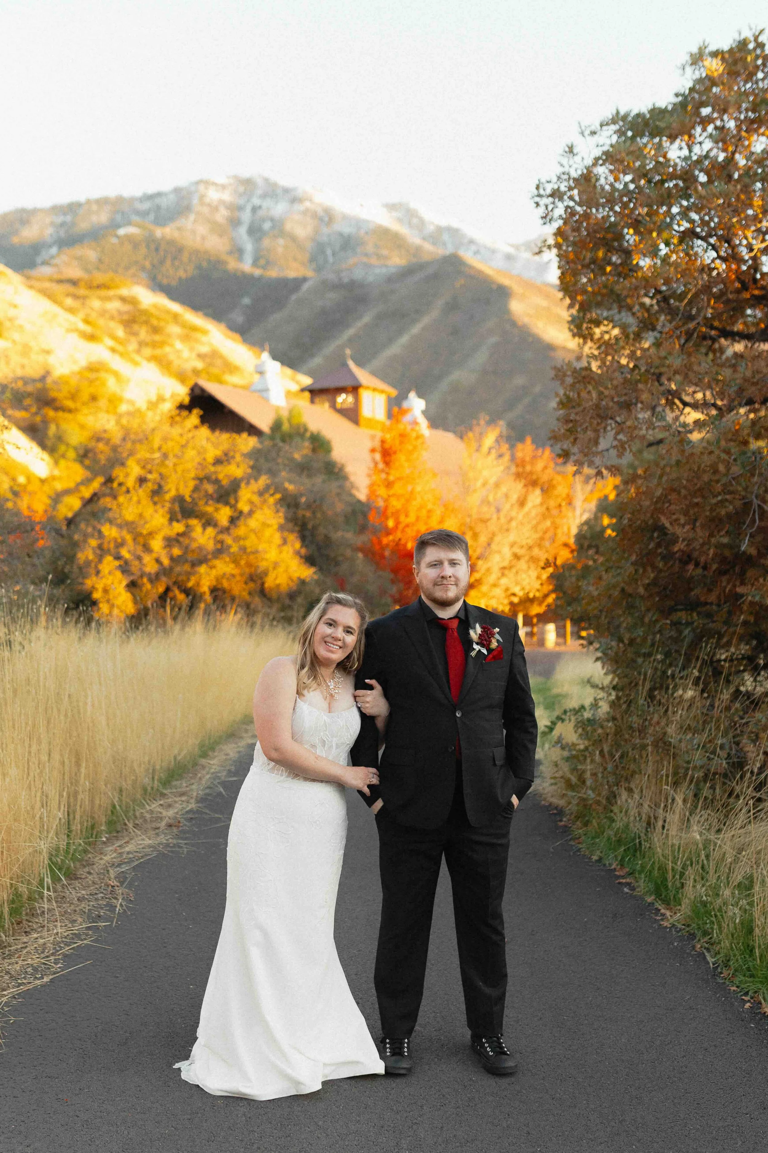 A bride holding onto her groom's arm and smiling in front of their wedding venue barn and mountain view in background.