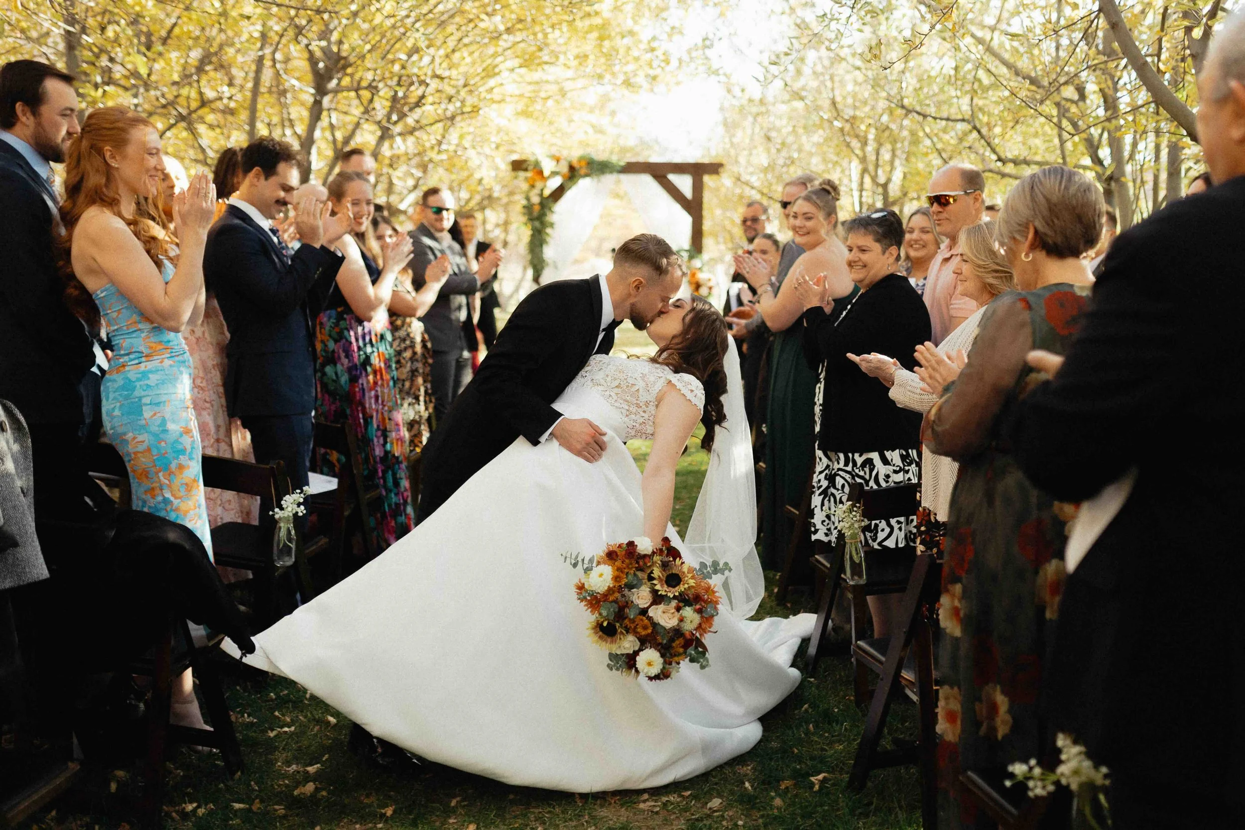 Bride and groom dipping after ceremony in an orchard of yellow fall trees in the background. Guests are applauding.