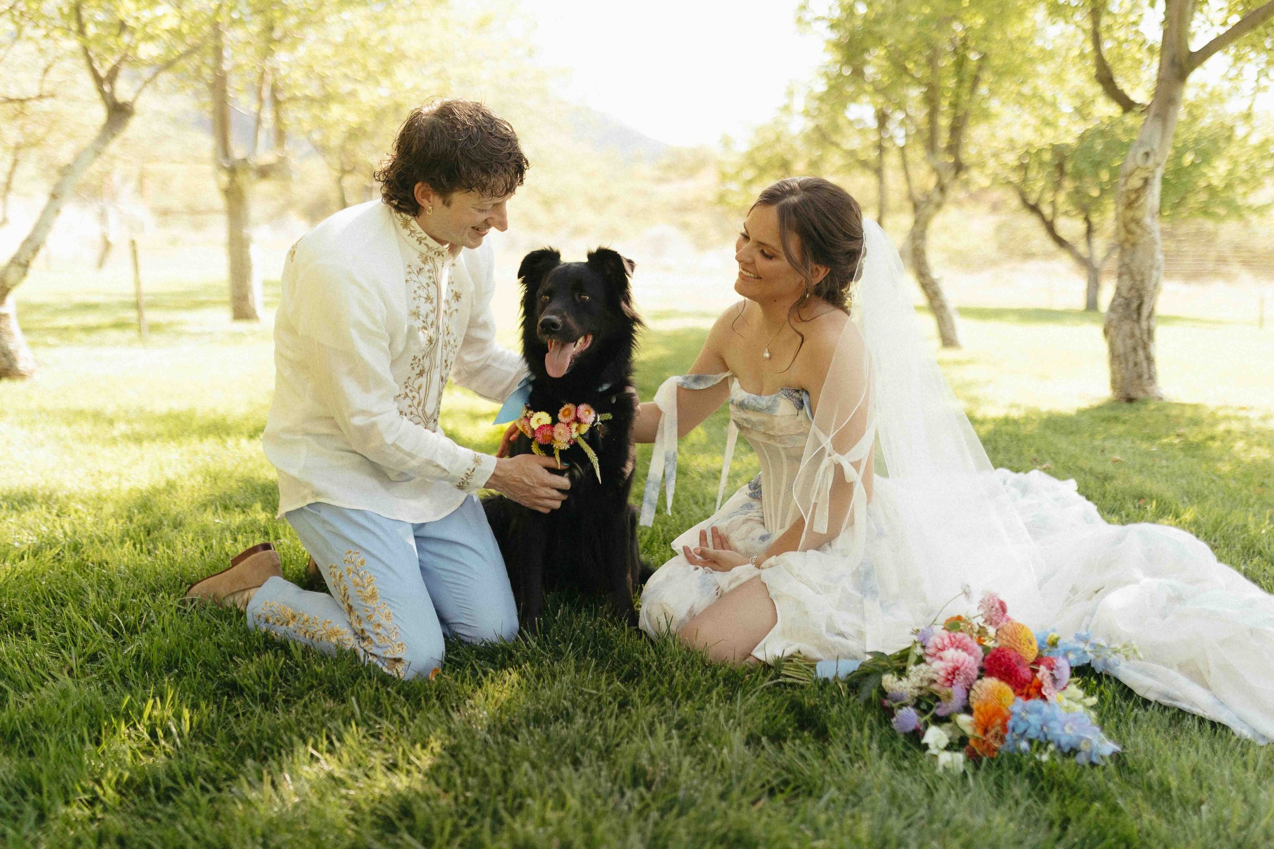 Bride and groom petting their dog in an orchard.