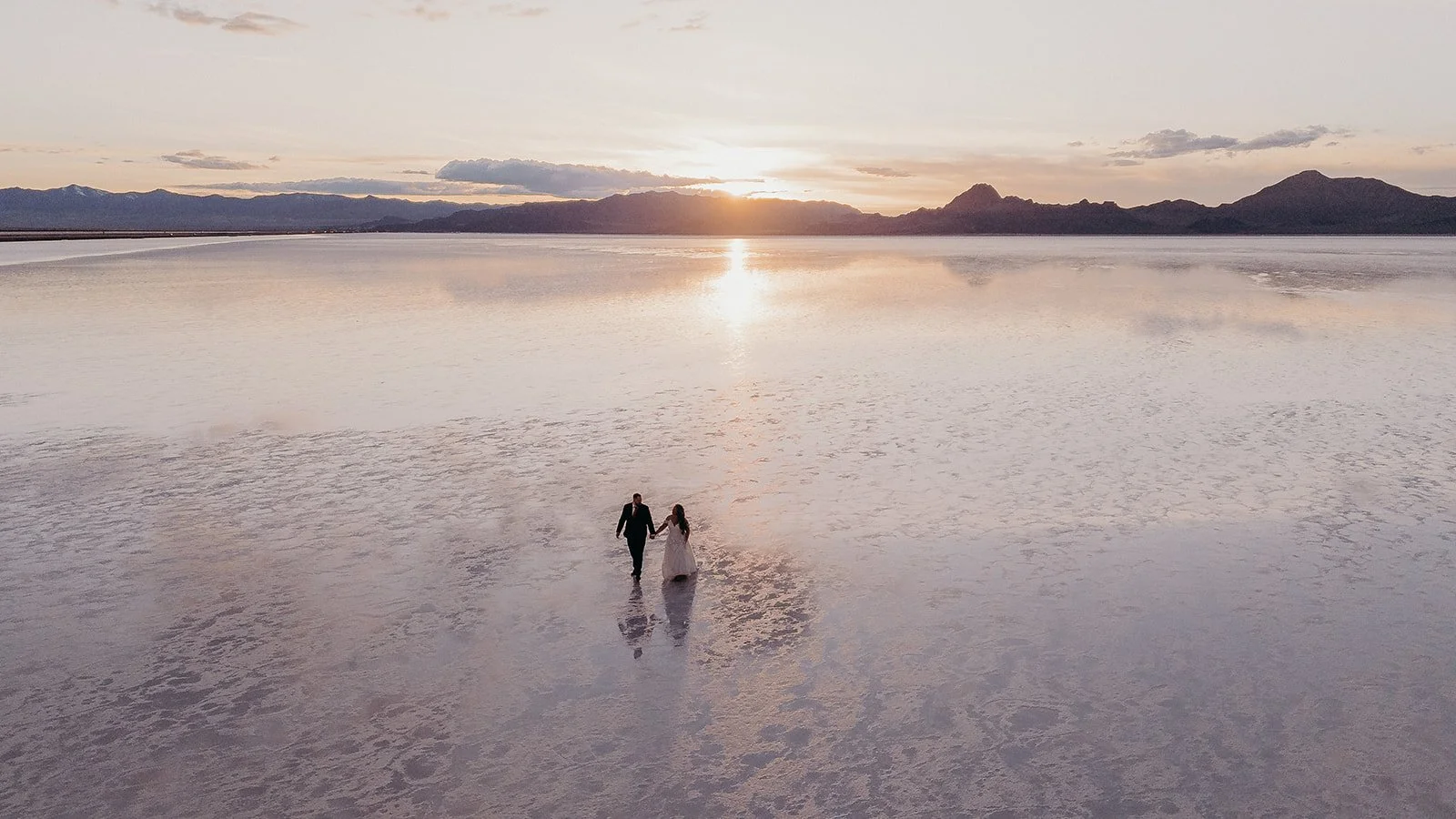 A couple in wedding attire holding hands and walking across a salt flat at sunset with mountains in the background.
