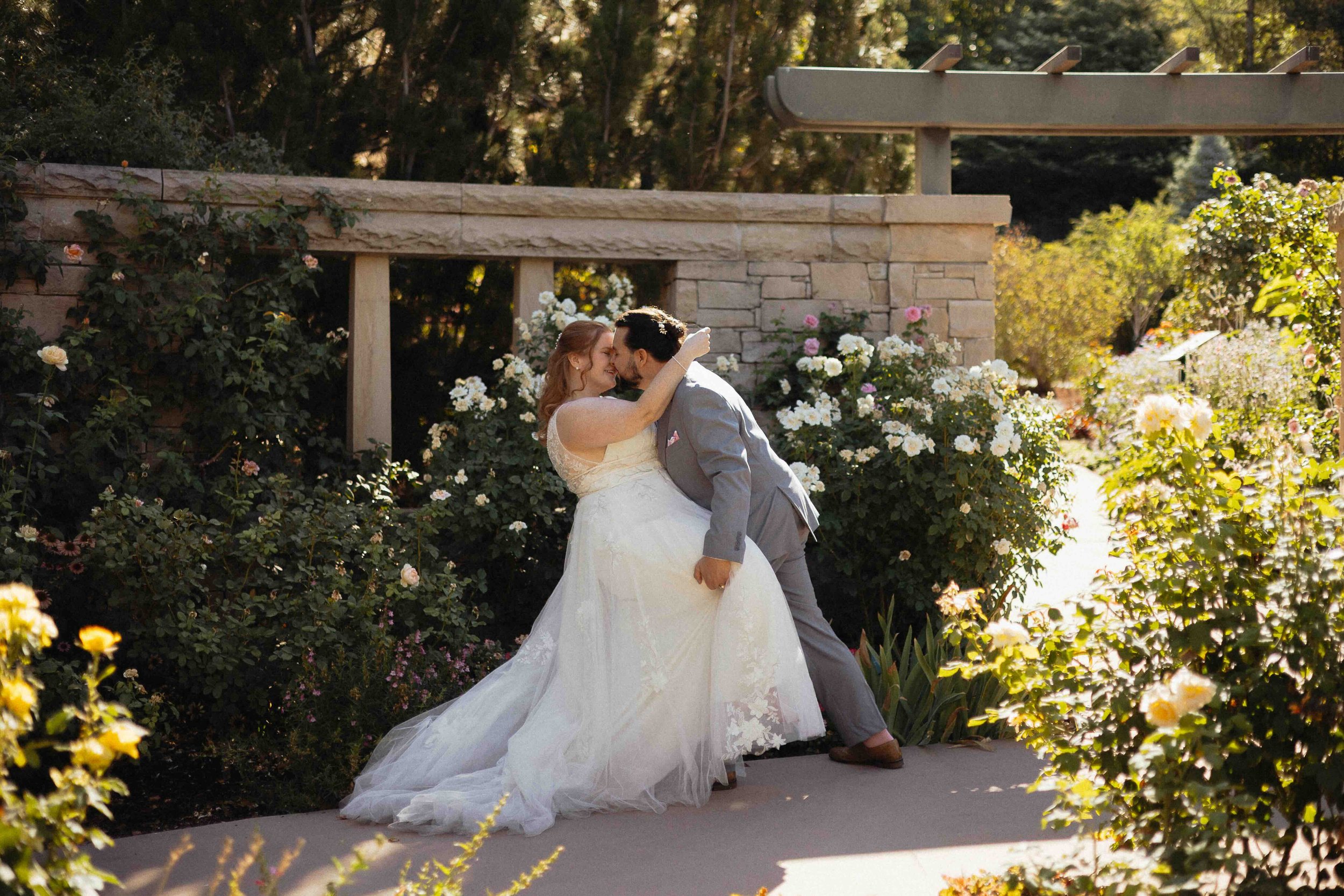 Bride and groom dipping in the Red Butte Gardens of Utah, with white roses in the background and a stone structure.