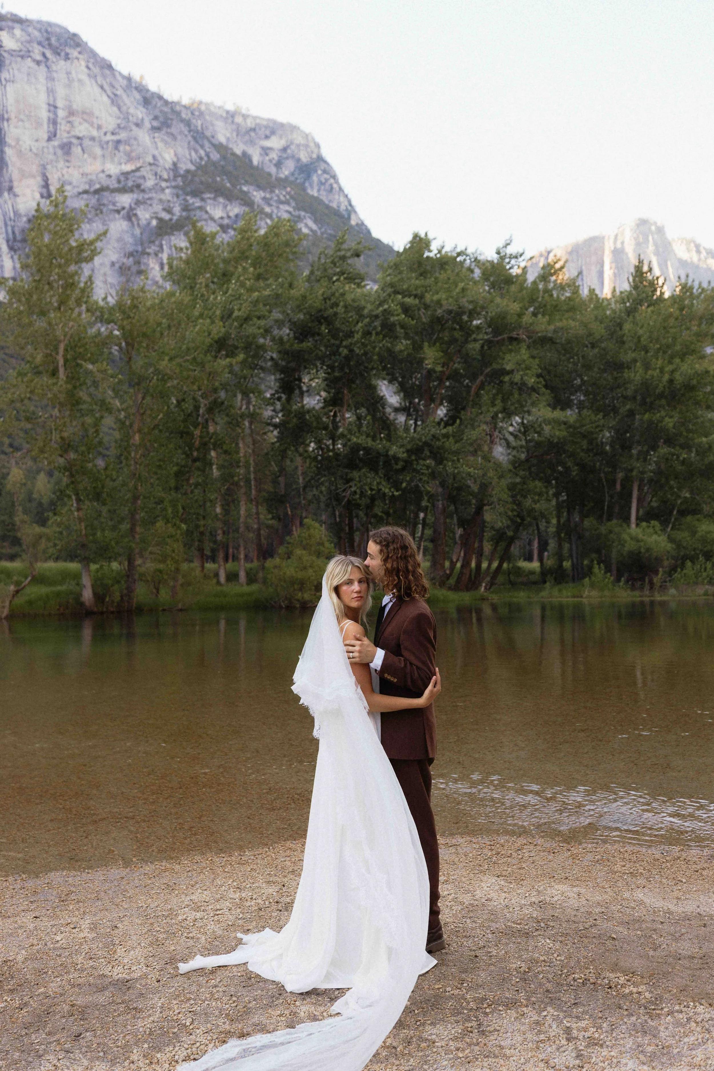 Bride and groom standing in front of the Merced River at Yosemite National Park with grey cliffs in the background.