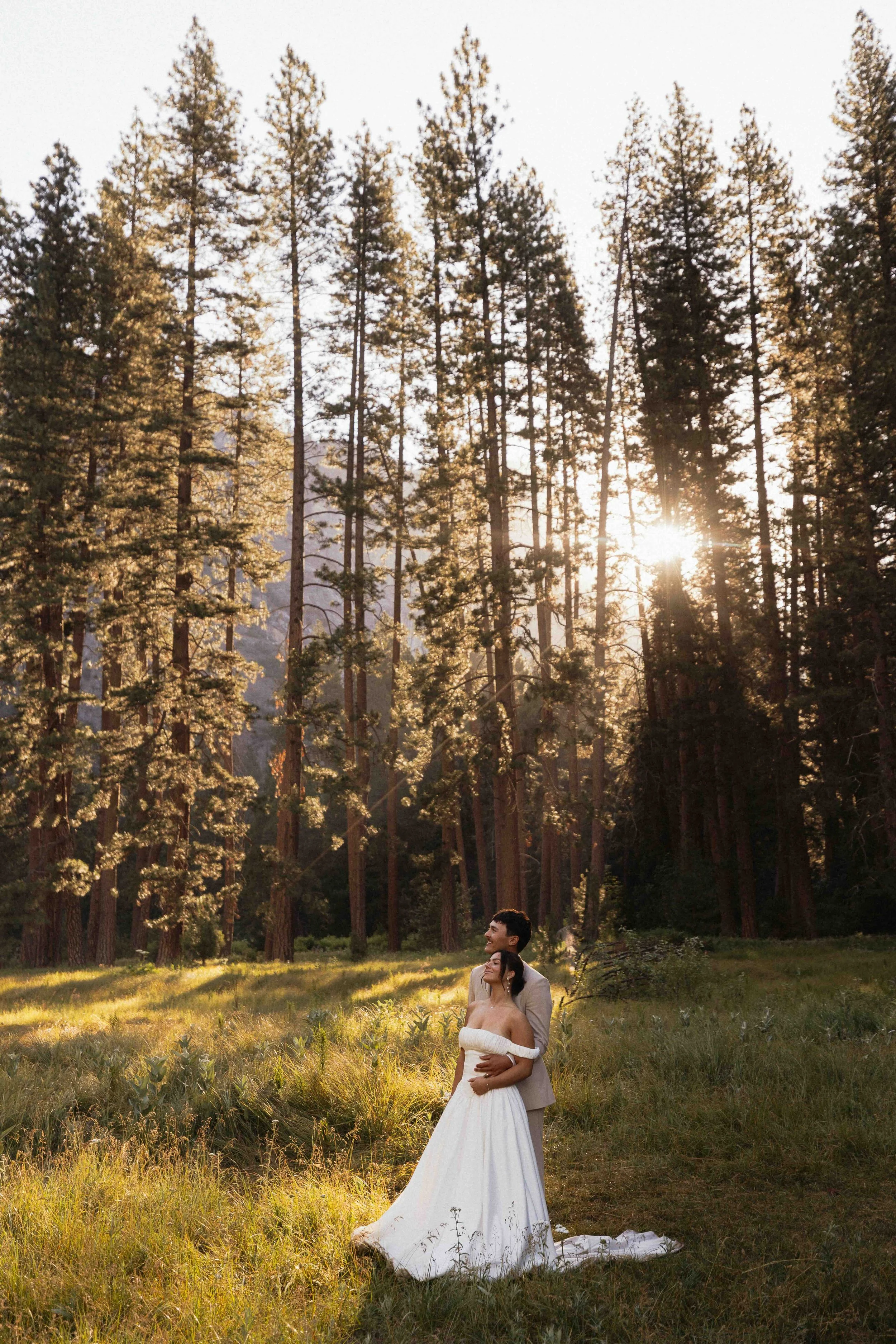 Bride and groom standing in front of sunrise and tall trees.