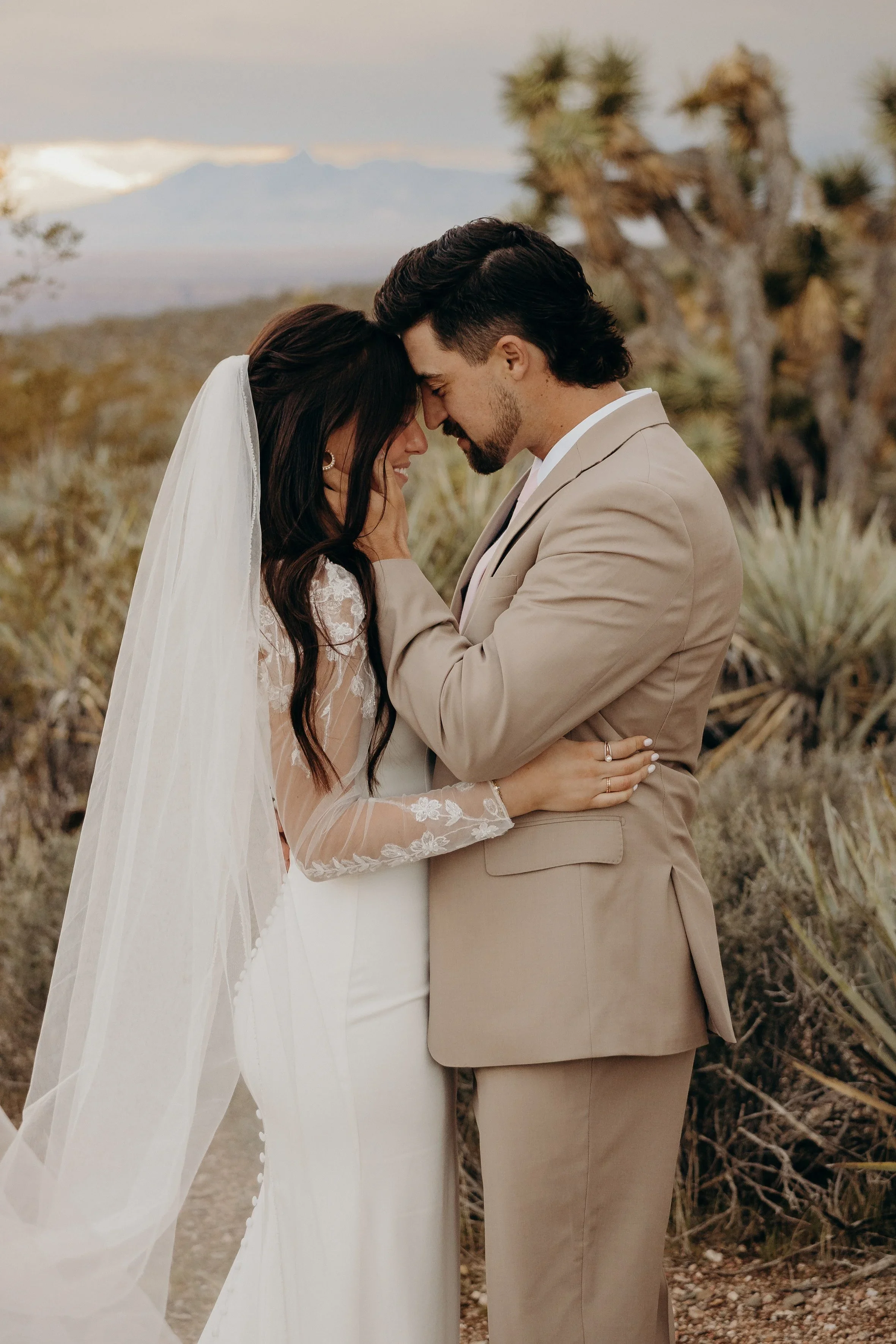 A bride and groom in an outdoor desert setting, touching foreheads and holding hands, with a bride in a white dress and veil and a groom in a tan suit, with desert plants and mountains in the background.