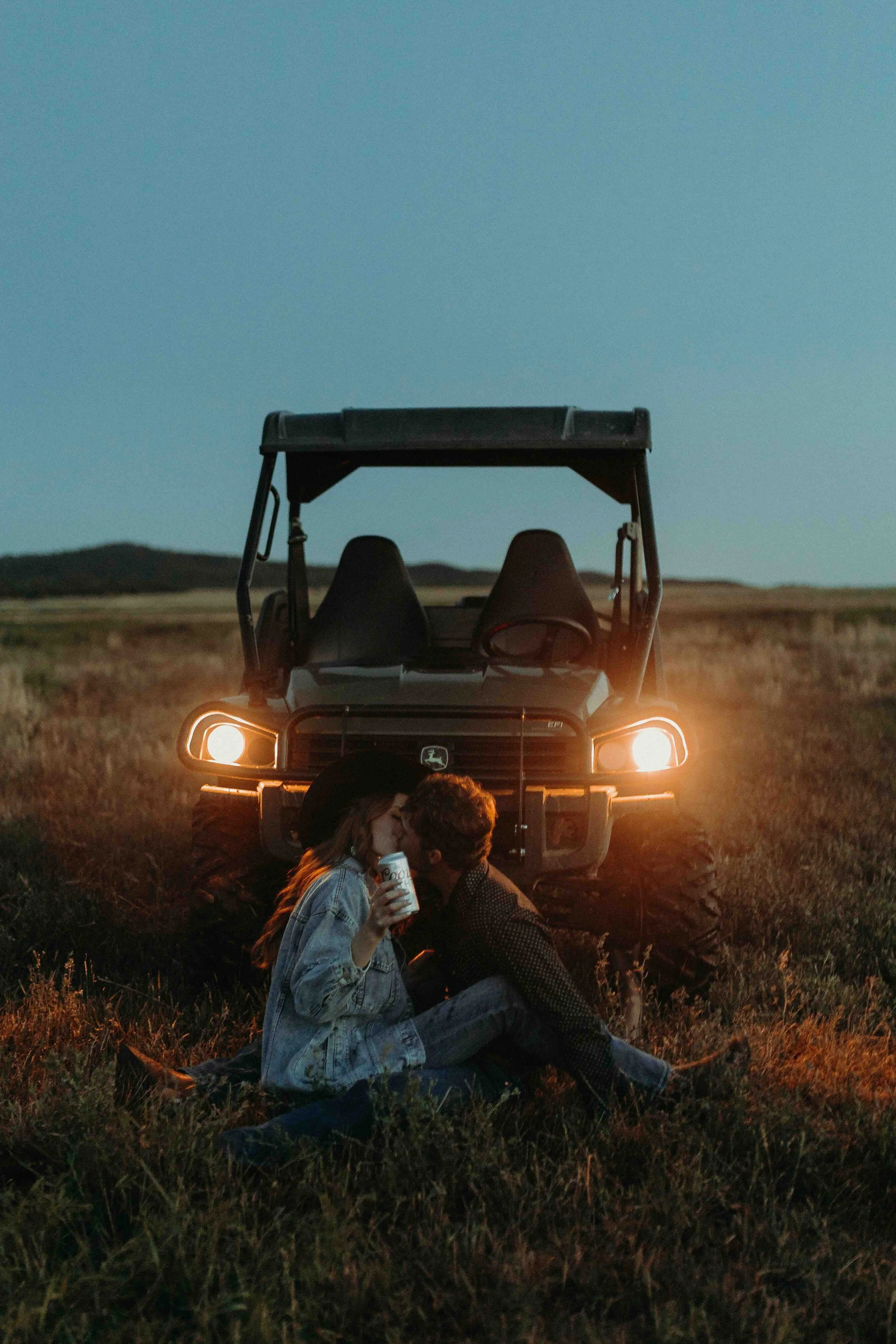 A couple sits on the grass in front of a John Deere utility vehicle during dusk, sharing a kiss. The woman holds a can while the vehicle’s headlights illuminate the scene, set against a rural landscape.