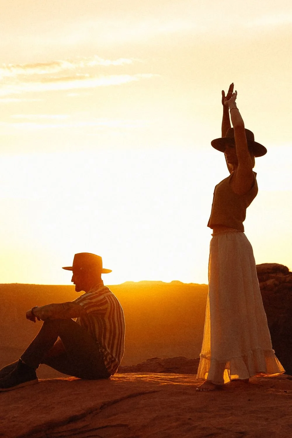 A moab desert couple. The man is sitting and the woman has her arms raised in the air soaking in the sunset.