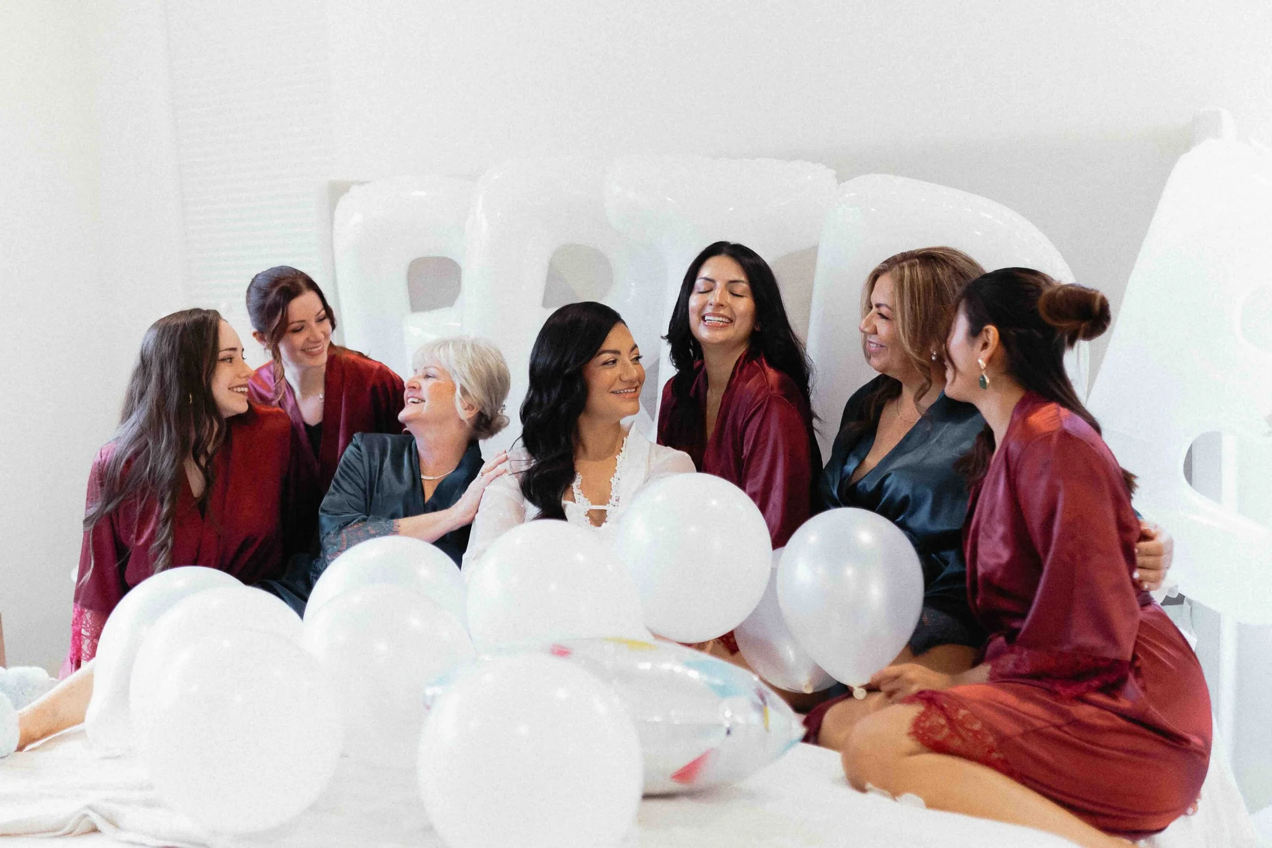 a bride surrounded by her bridesmaids in robes on a bed with balloons.