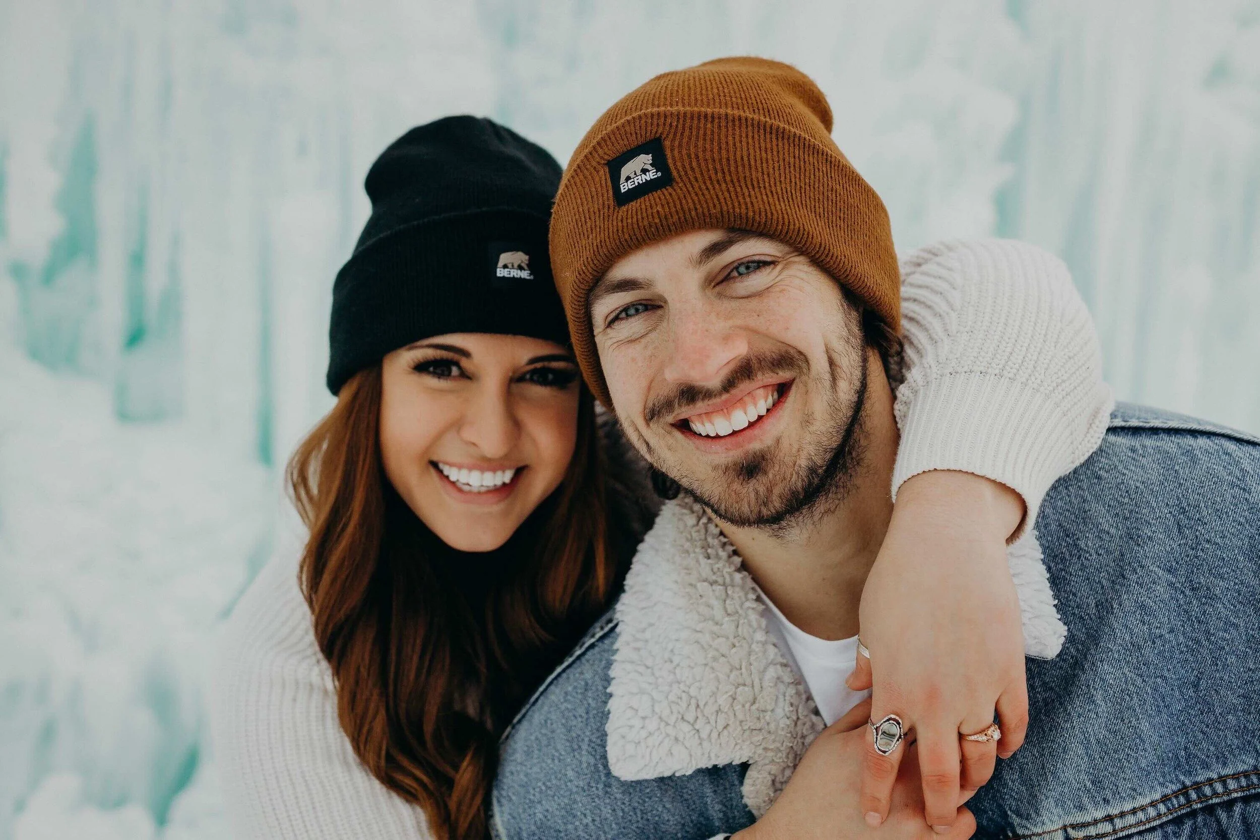 A smiling man and woman in winter clothing, wearing beanies and jackets, enjoying cold weather outdoors with snow and trees in the background.