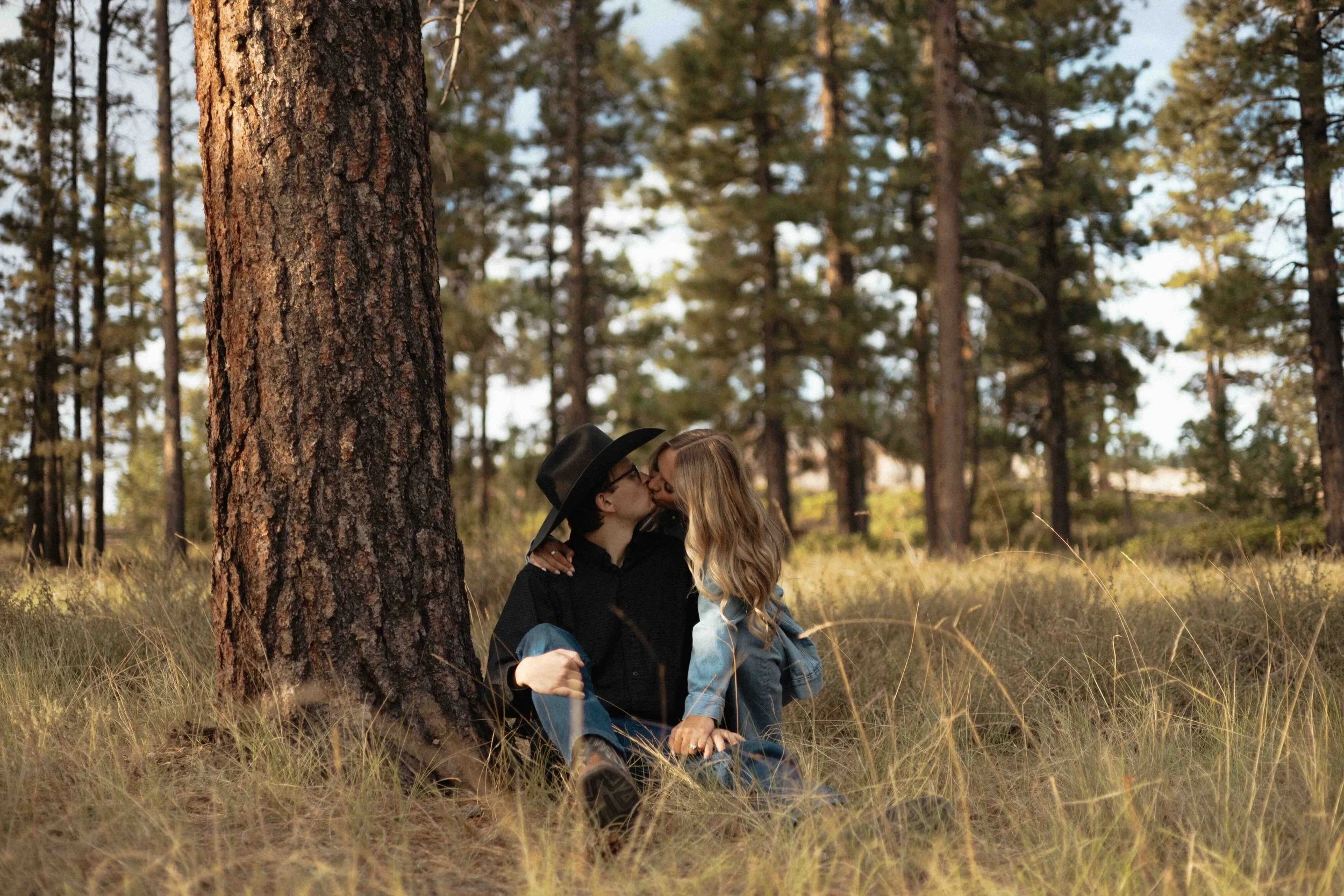 A couple sitting in the grass kissing, leaning against a tree in a forest with tall trees and sunlight.