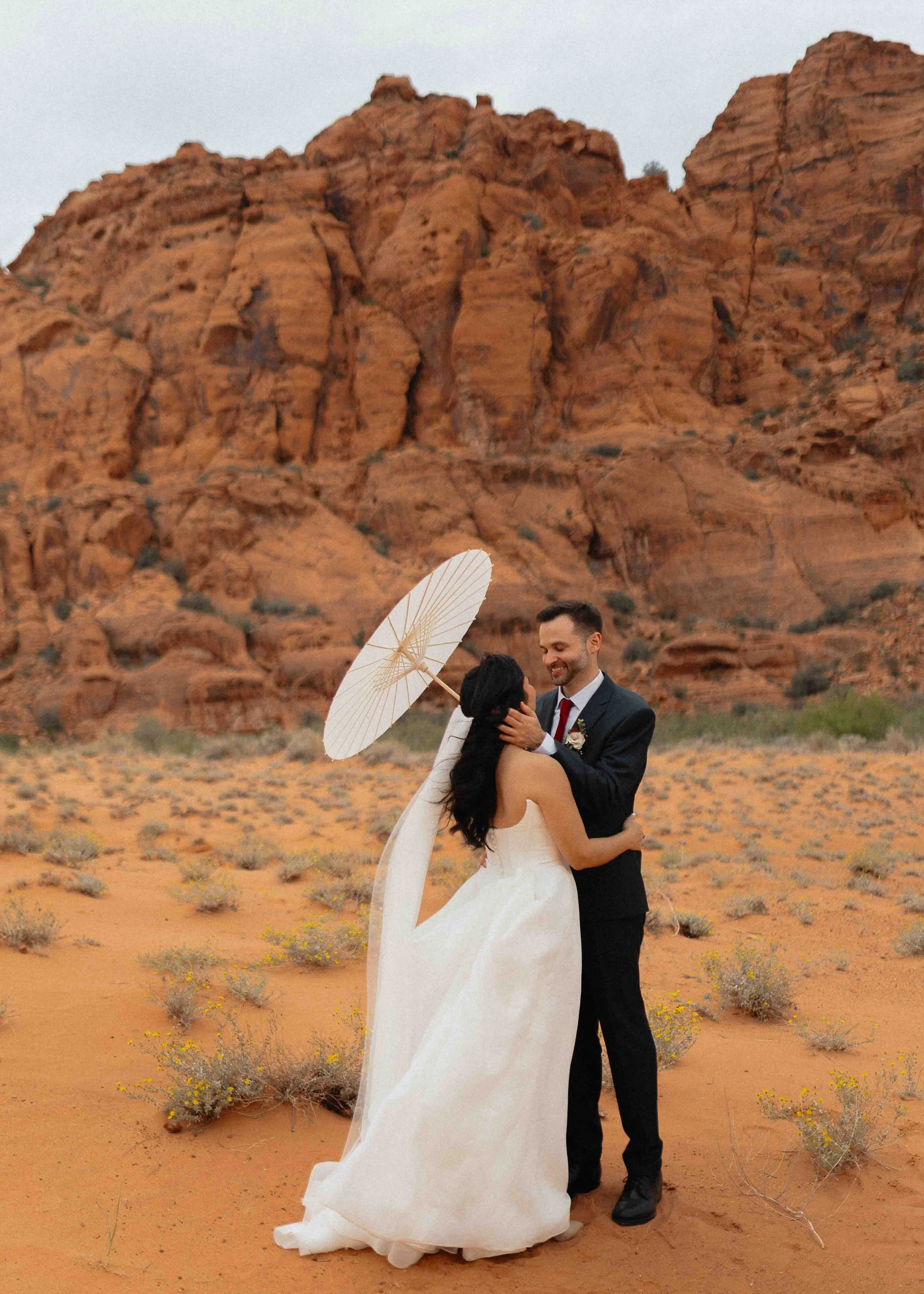 A bride and groom kissing with a white parasol in front of rocky formations in the background.