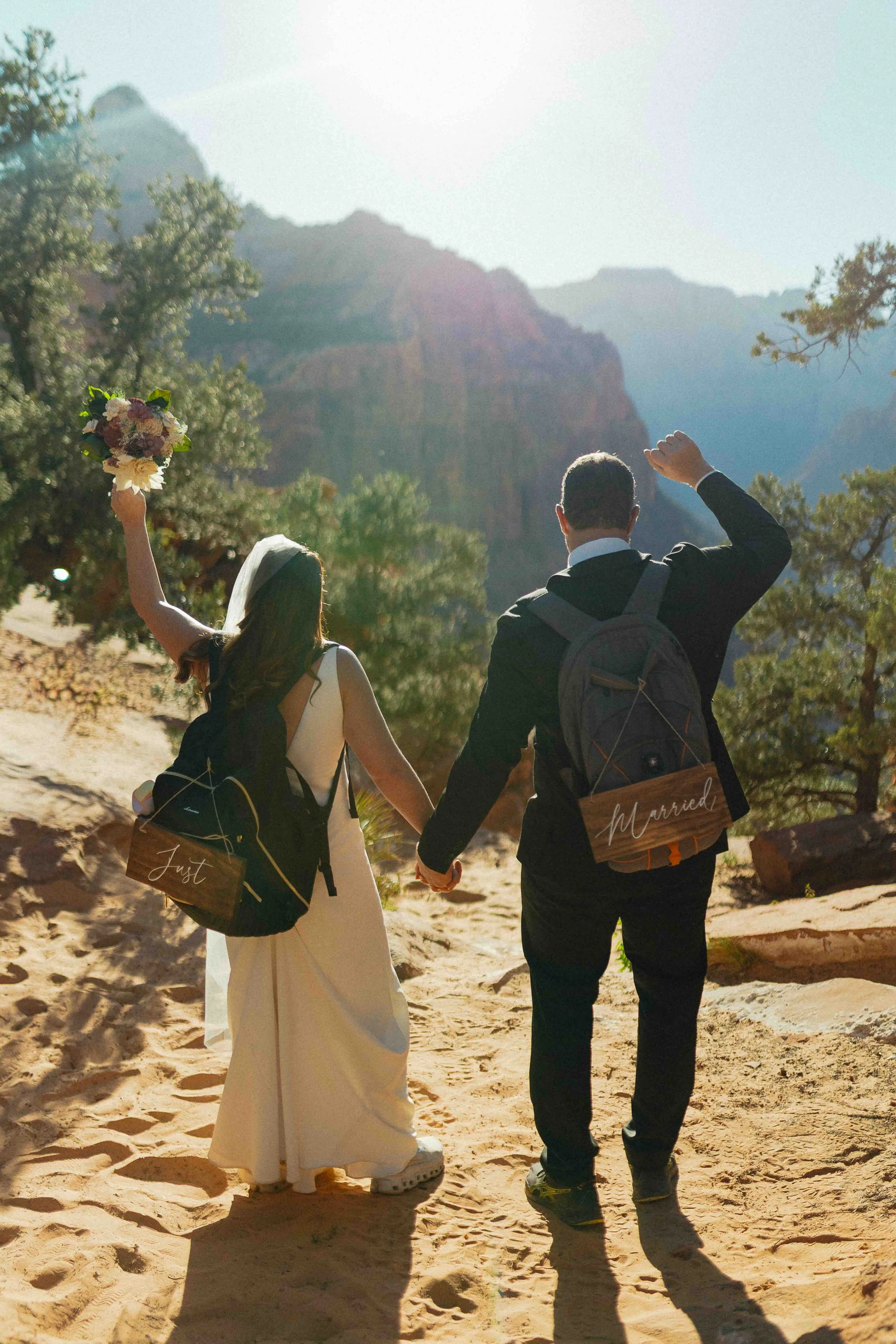 Couple wearing backpacks with "Just Married" signs on the back. Their hands are waving in the air.