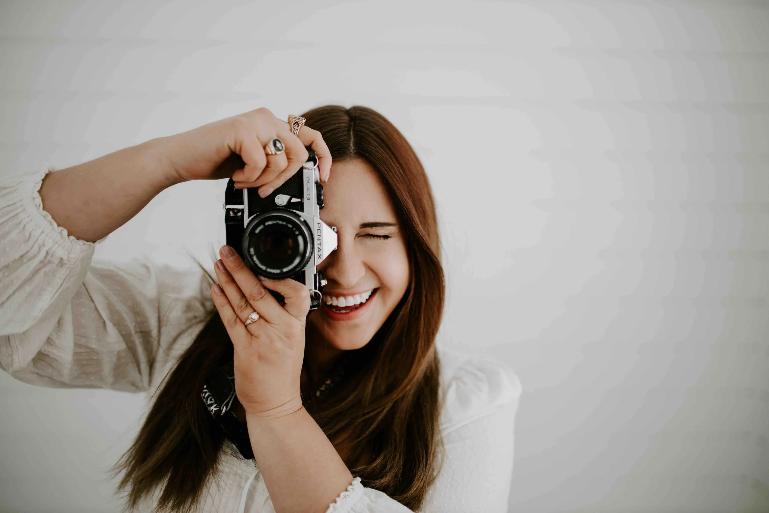 A woman with long brown hair taking a photo with a vintage camera, smiling with her eyes closed, against a plain white background.