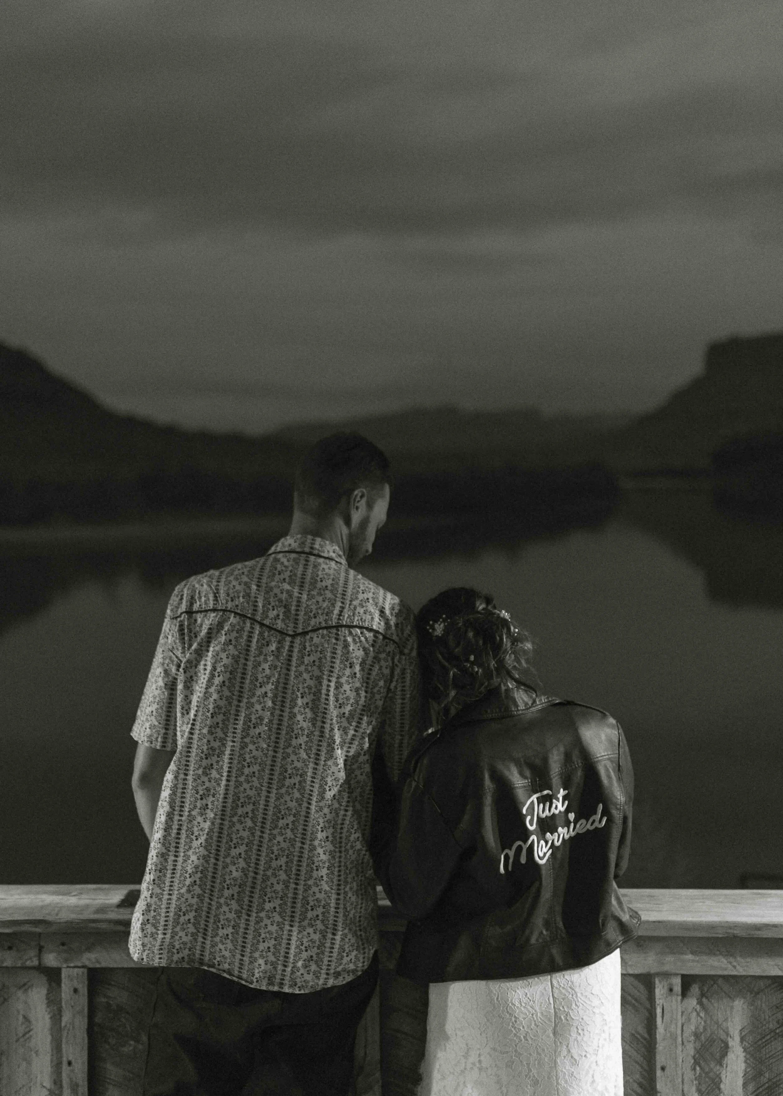 Bride in a leather jacket that says "Just Married"  and leaning on her groom while overlooking the river at night.