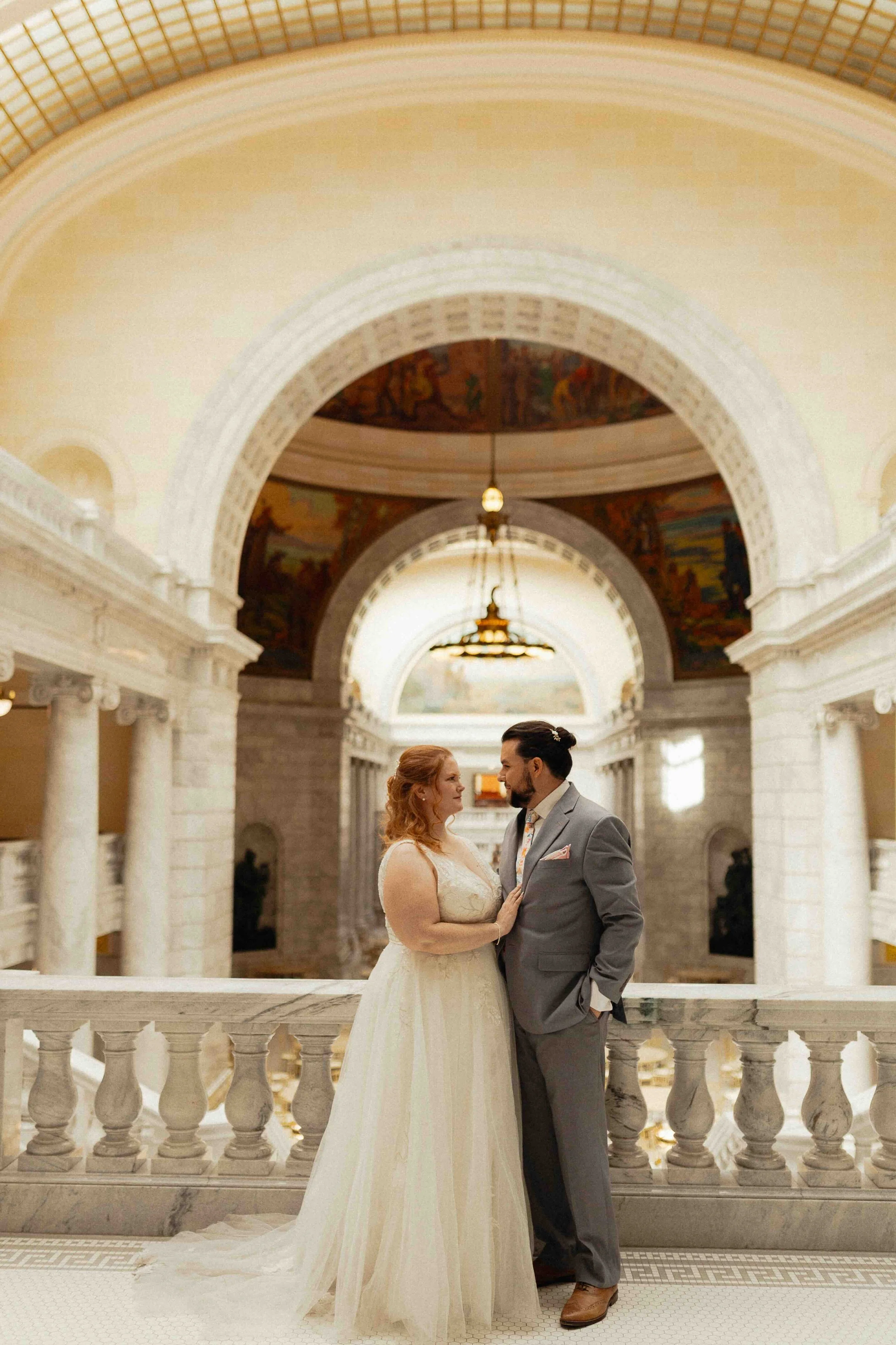 Bride and groom holding onto one another in front of the inside of the Utah State Capitol Building with a domed ceiling and chandelier in the background.