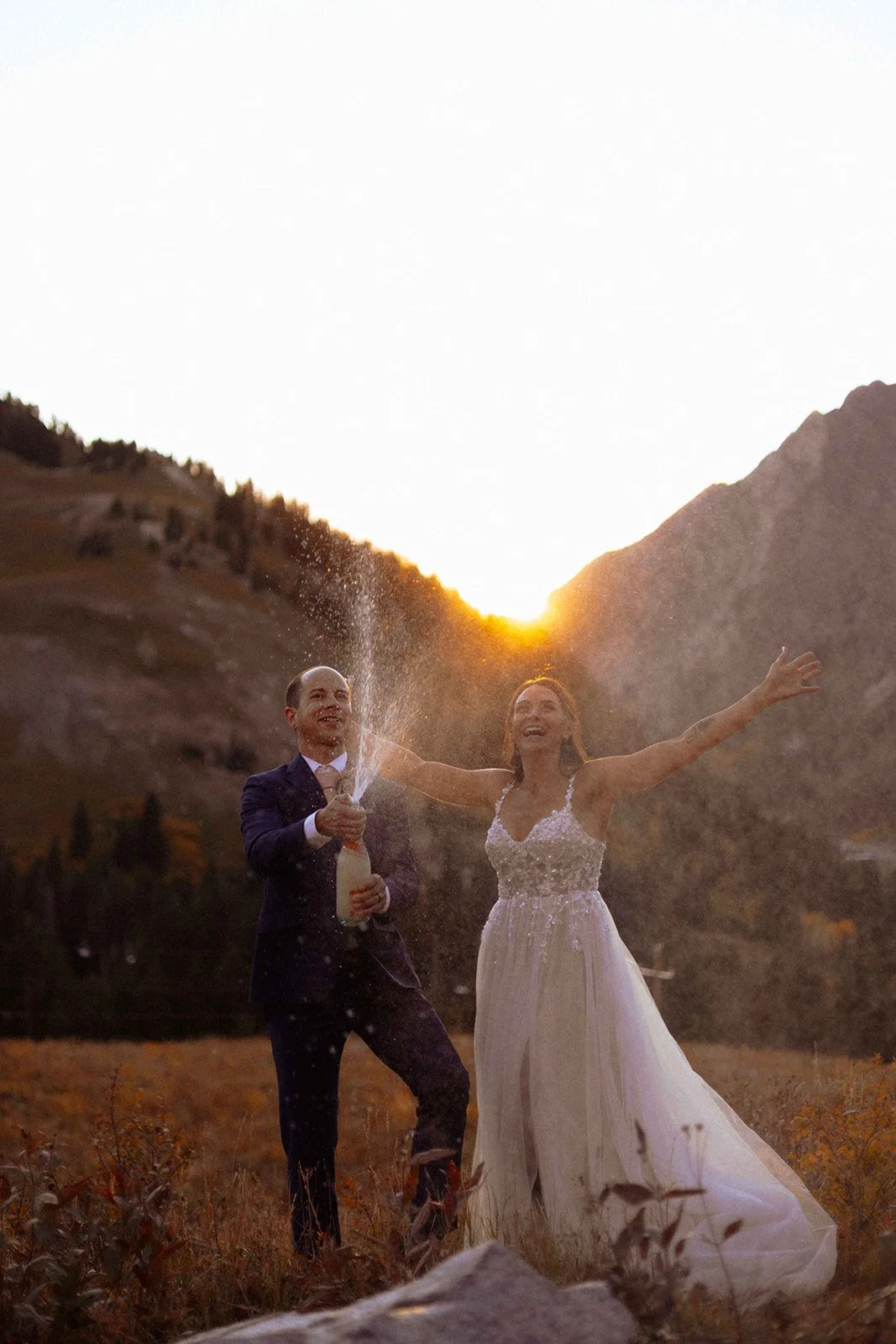 A newlywed couple celebrating outdoors at sunset, with the groom in a dark suit and the bride in a white wedding dress, spraying champagne in a mountainous landscape.