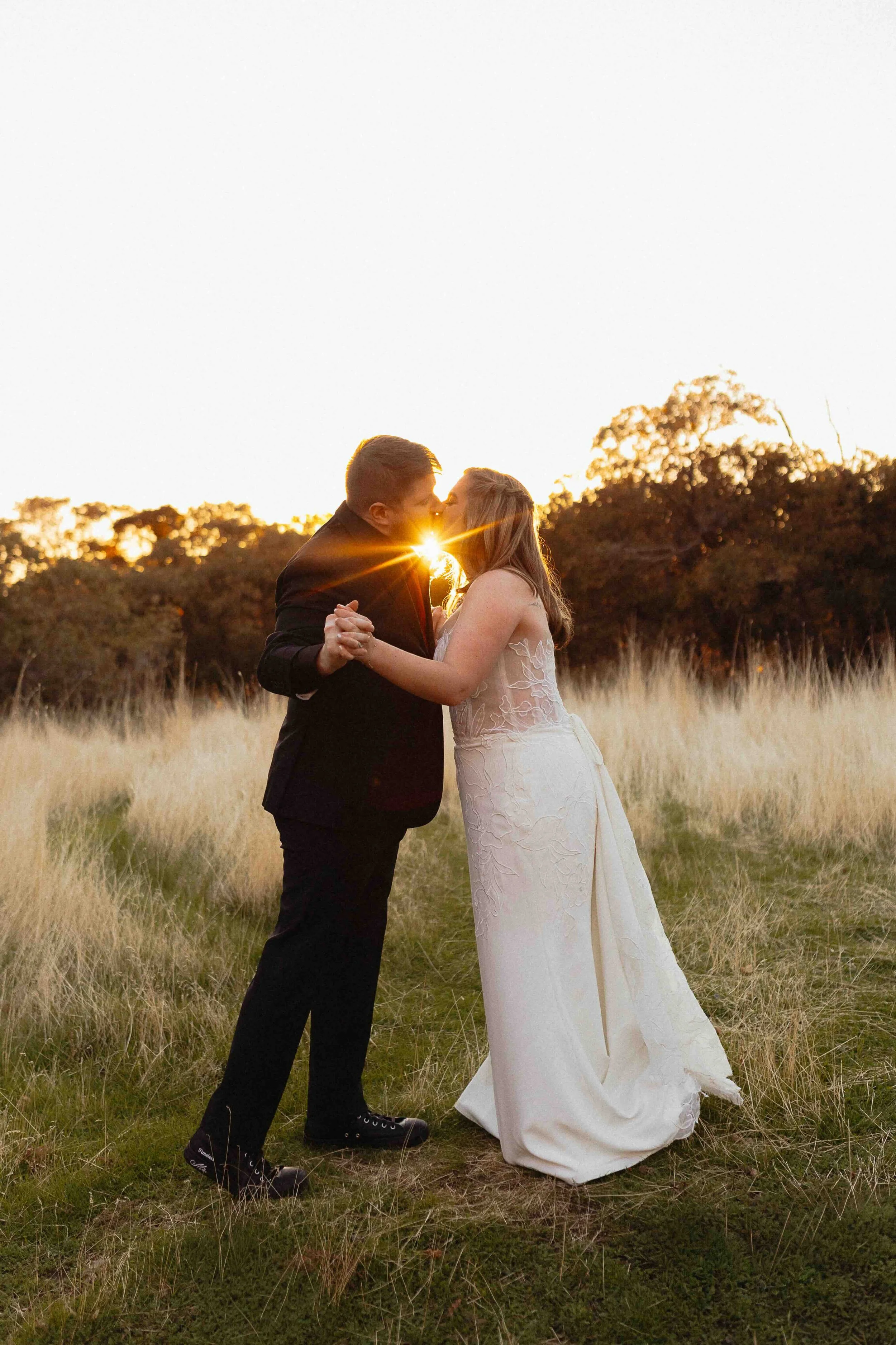 Bride and groom kissing in field with long grasses. The sunset is streaming through their kiss.