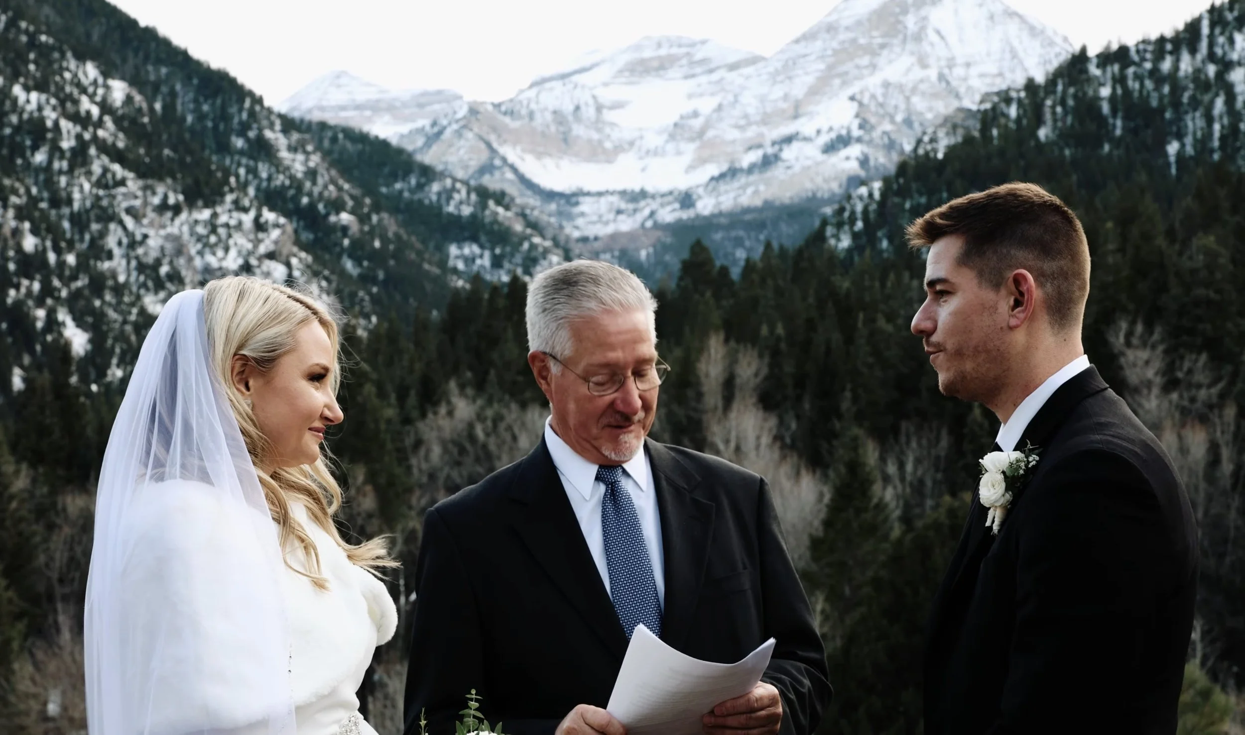 Bride, groom, and officiant in front of snowy mountaintop.