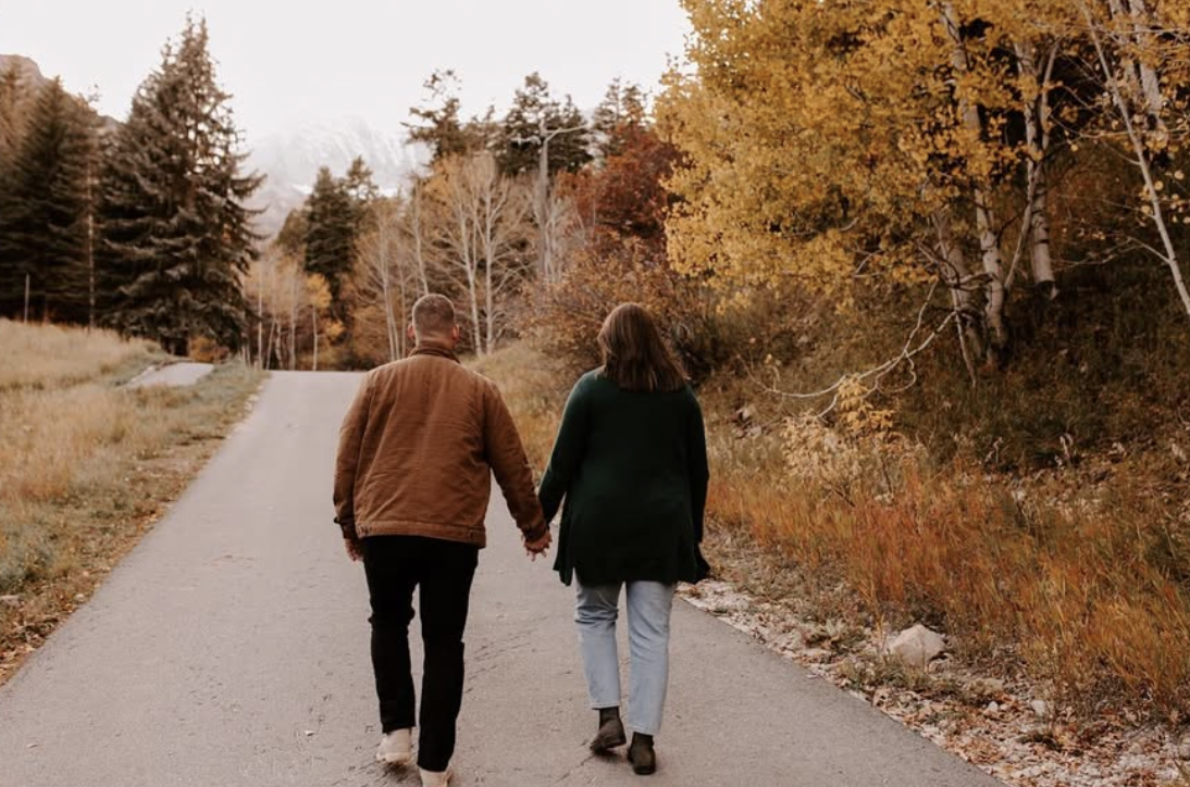 Couple holding hands and walking up path with fall trees in the background.