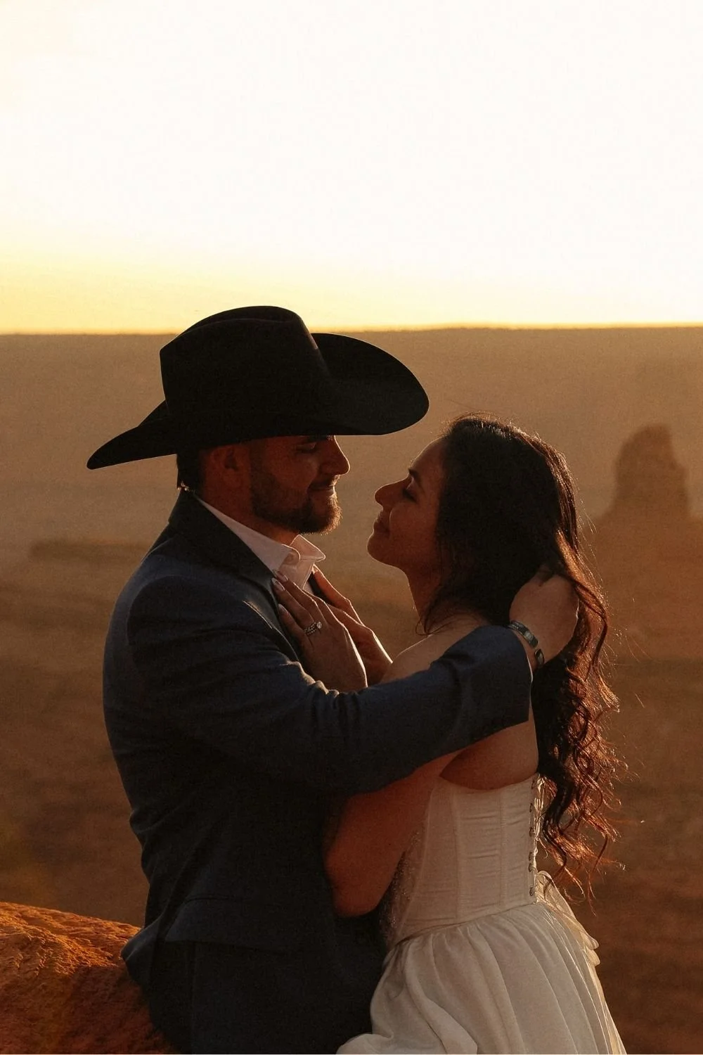Groom in cowboy hat brushing back bride's hair in front of a Moab red cliff sunset.
