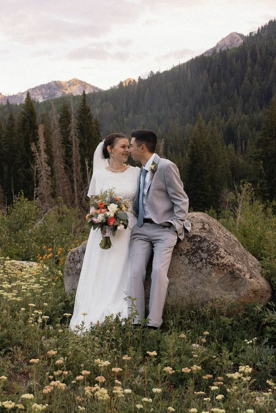Bridal couple about to kiss in flower field with mountains in the background.