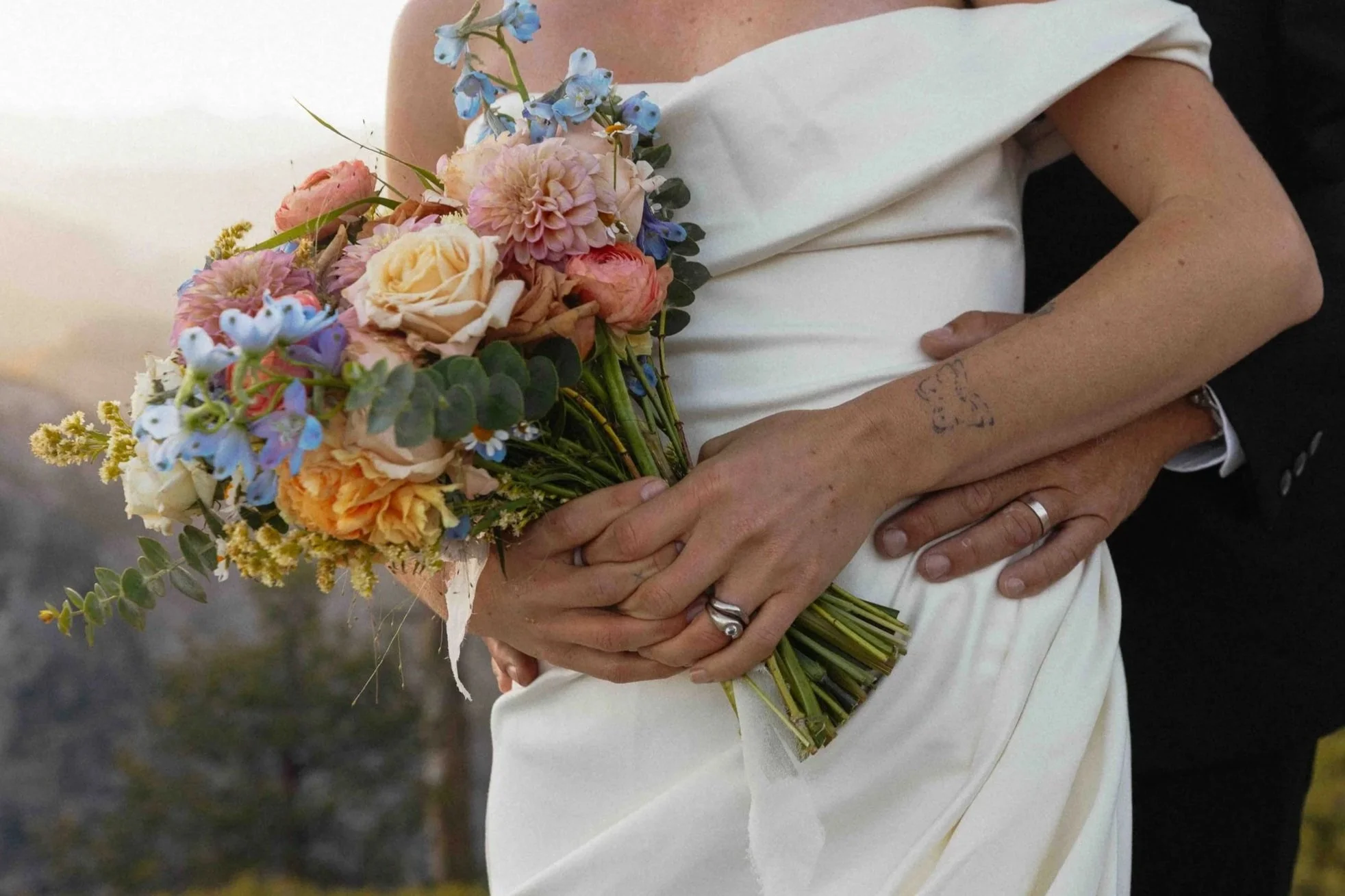 Bride and groom hugging around each other. The bride is holding her bouquet and there are cliffs in the background.