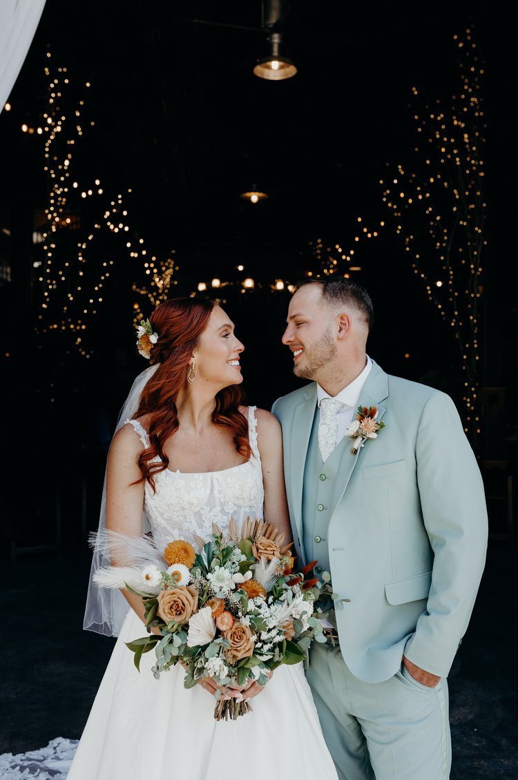 Bride and groom smiling at one another with a dark background with twinkly lights shining.