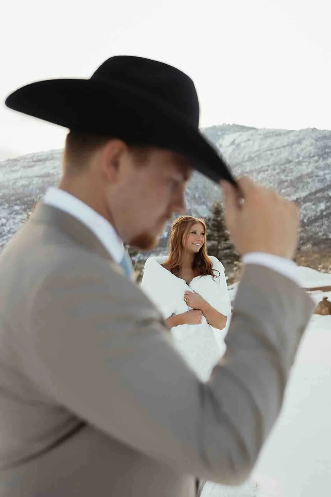 Groom holding onto his cowboy hat and bride looking off into the distance in a snowy mountain Utah landscape.