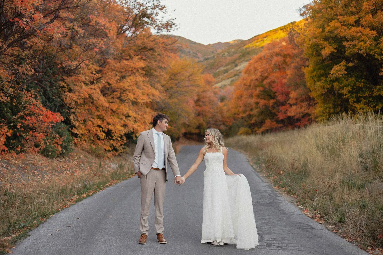 A bride and groom holding hands on a paved road surrounded by colorful autumn foliage.