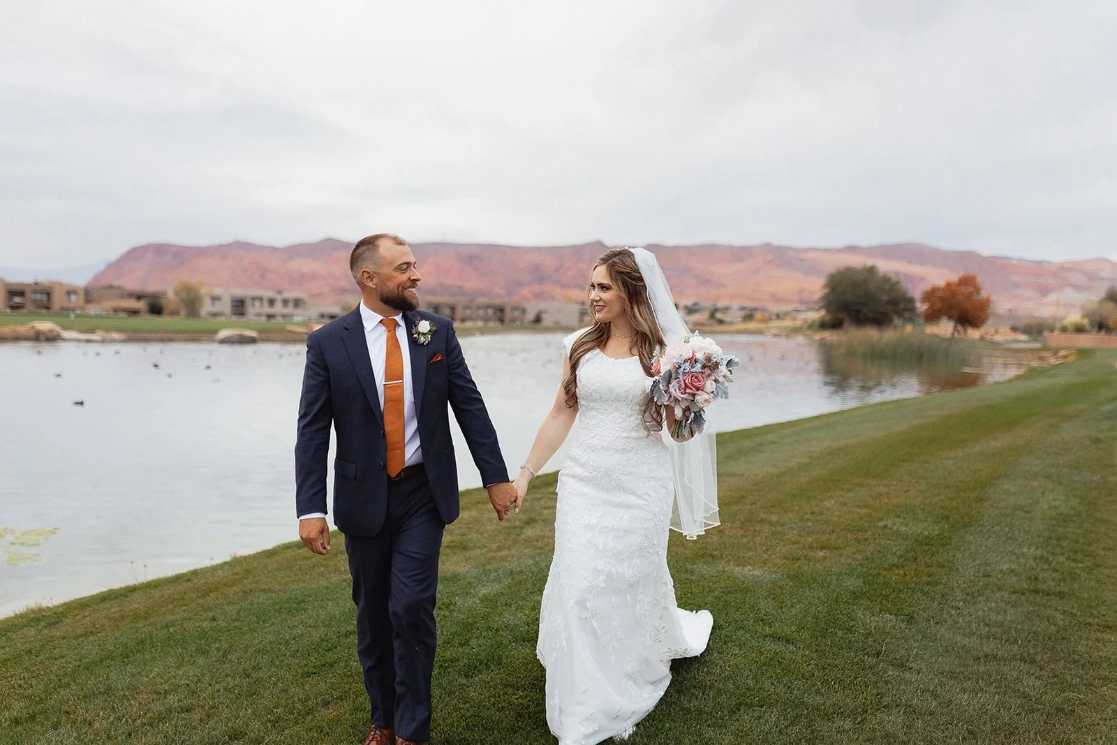 Couple holding hands in front of golf course pond with red rock cliffs in the background.