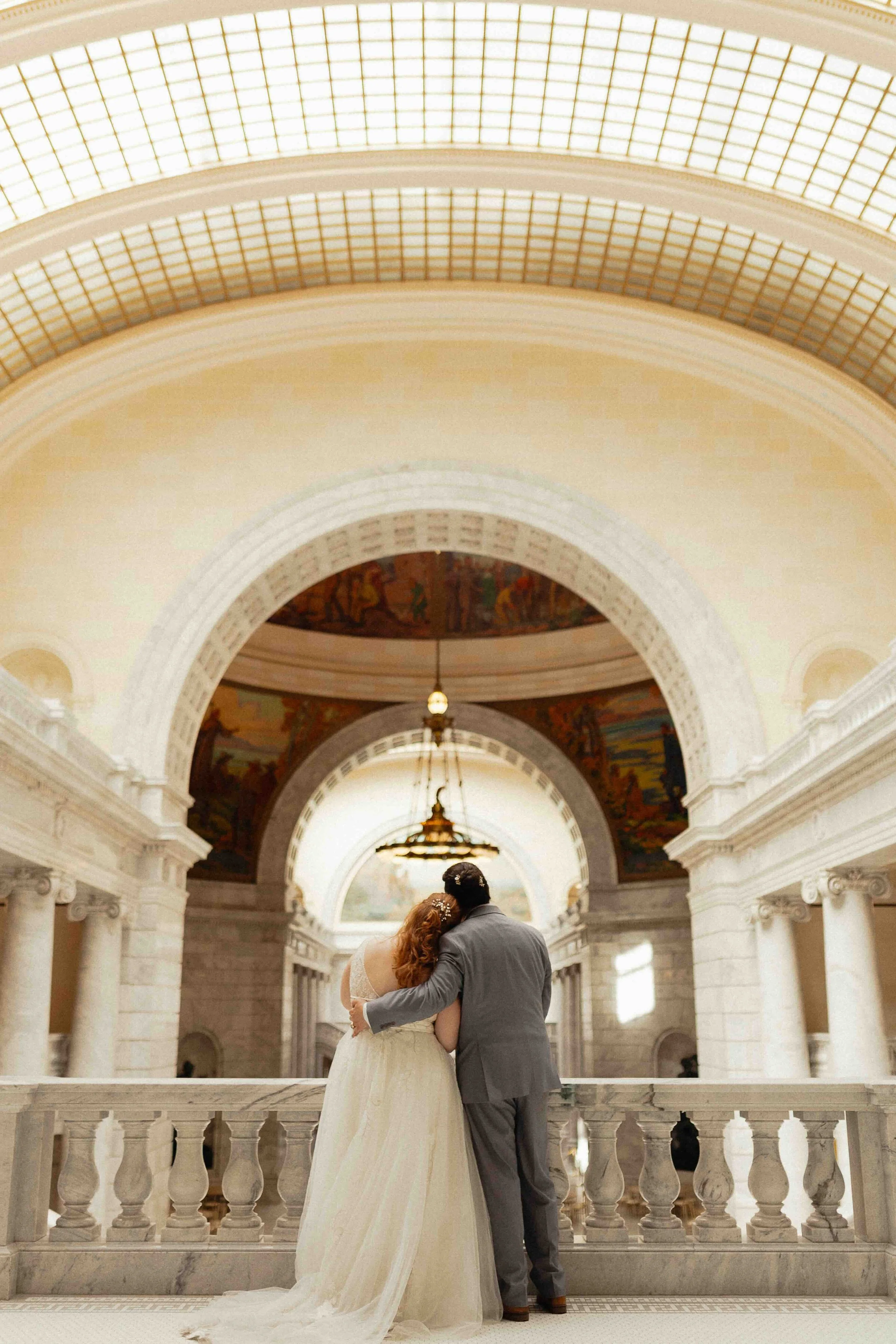 Bride and groom holding onto each other and looking out at the domed ceiling of the Utah state Capitol building's interior.