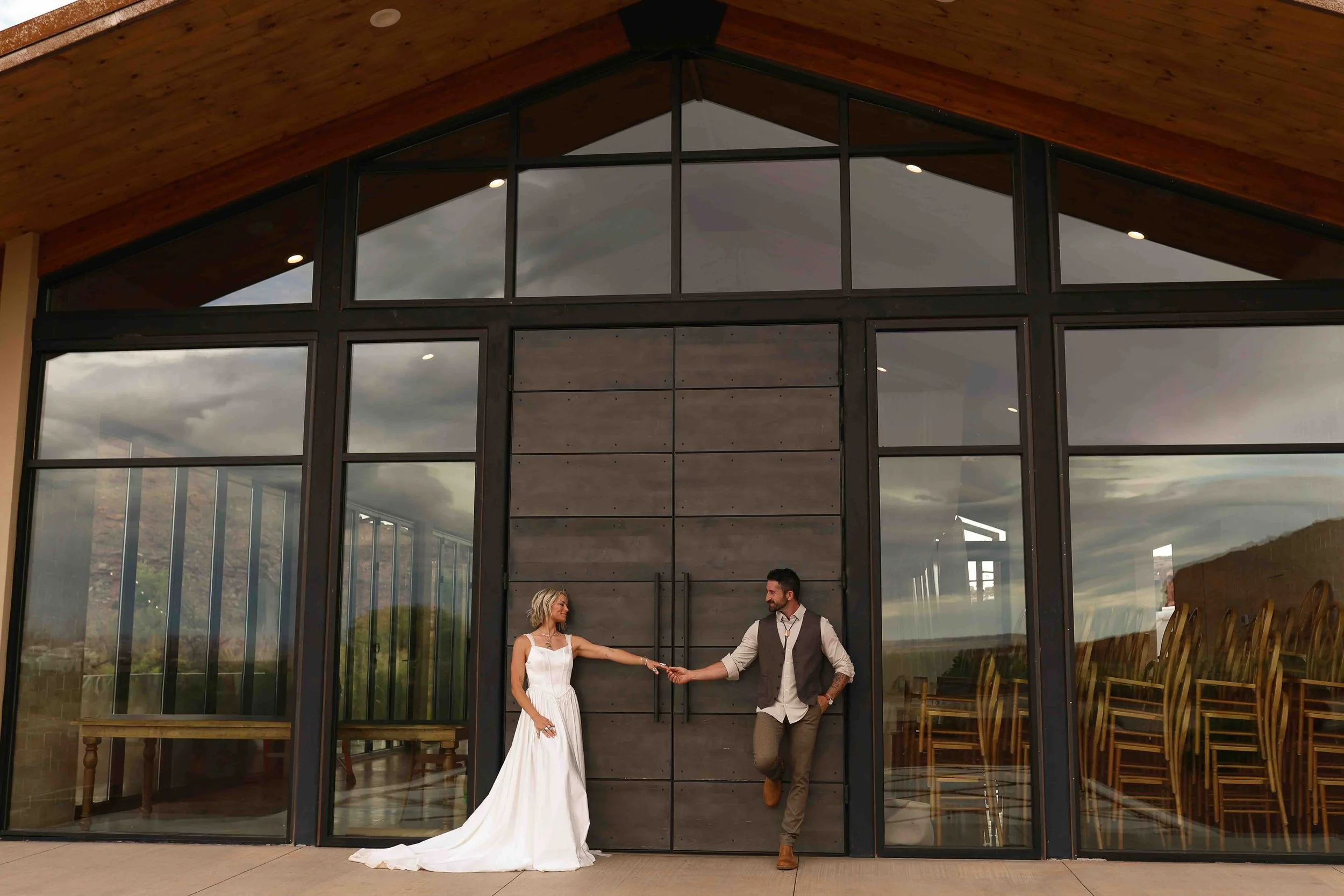a bride and groom holding hands in front of the red earth venue in Moab, UT. It is an A-frame building with windows on the outside.