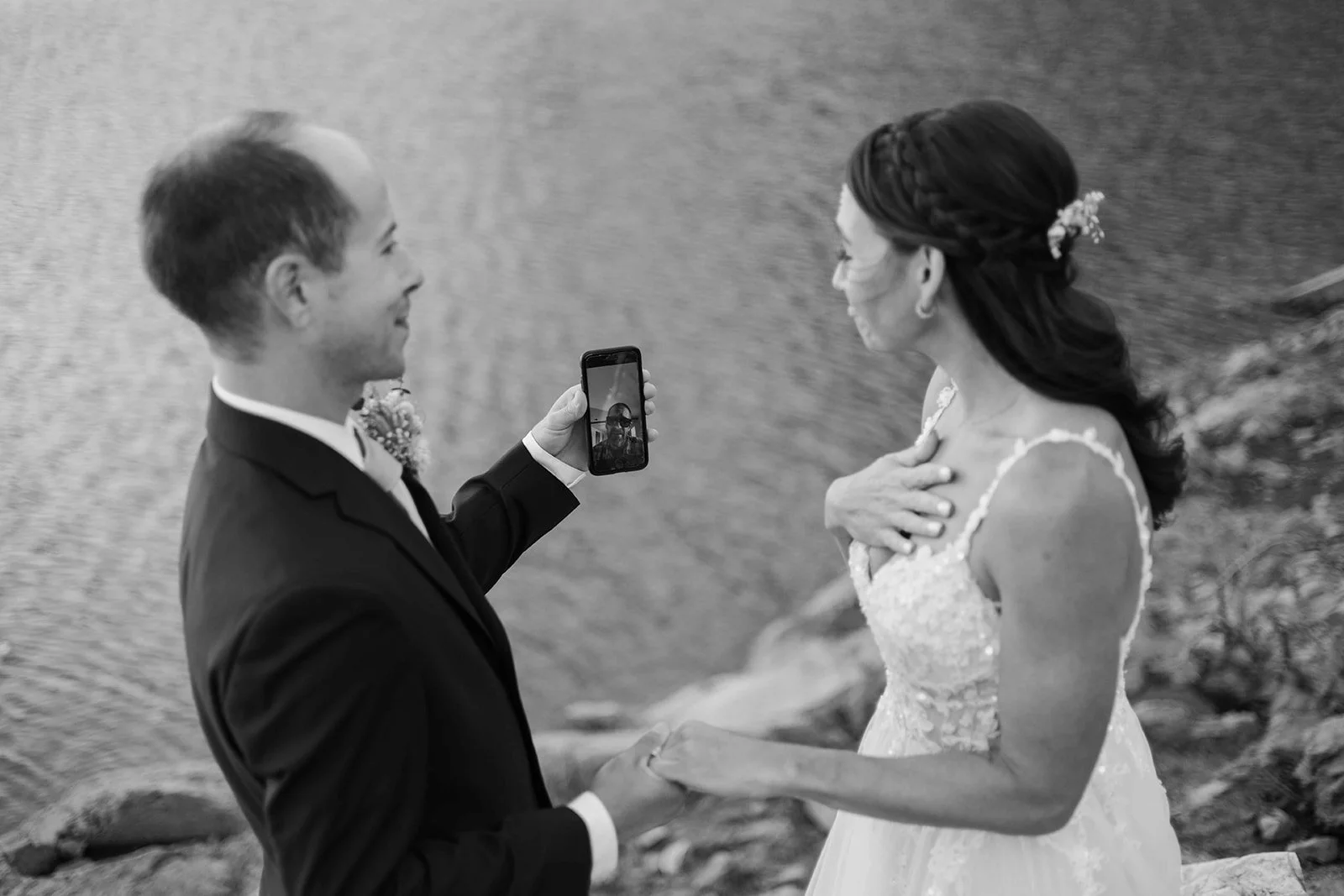 Bride and groom watching a video on their phone in front of a lake.
