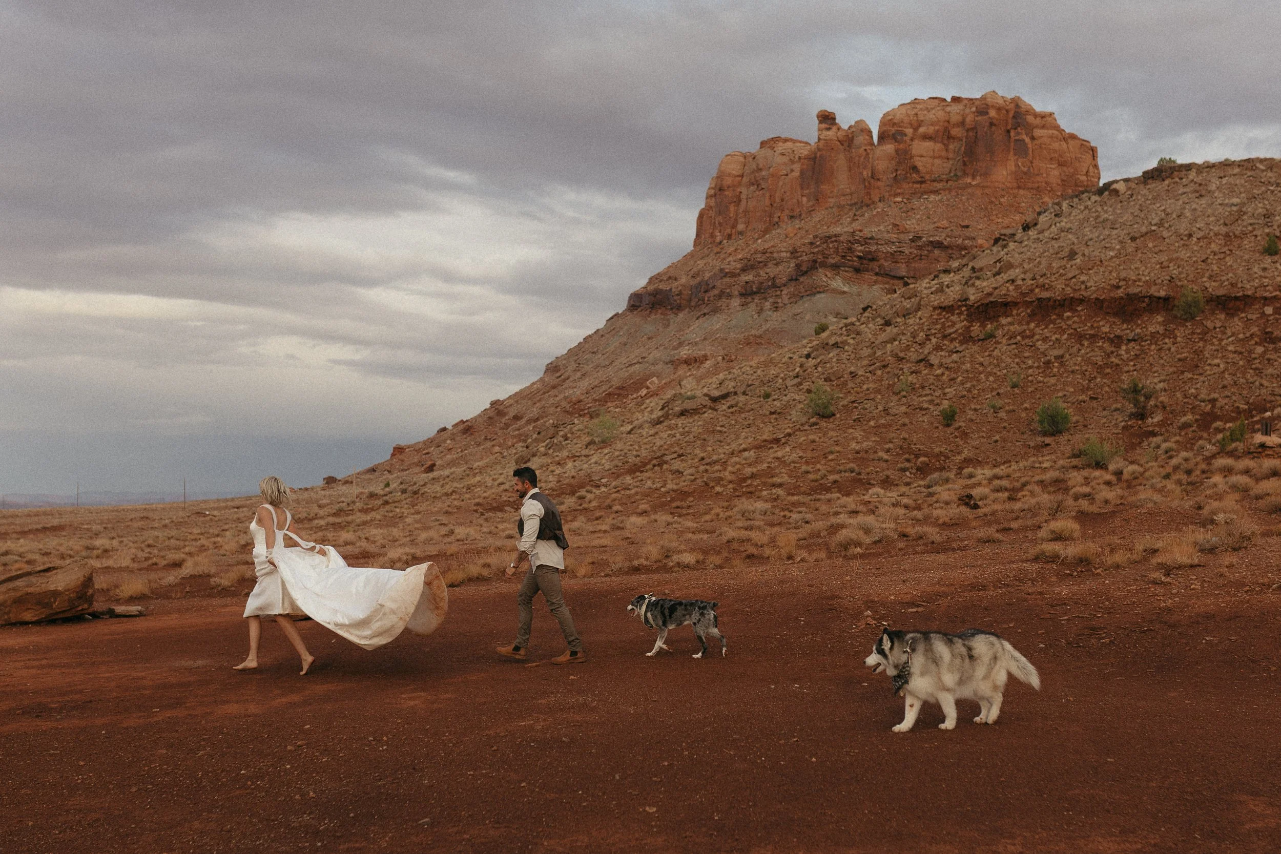 A woman in a white dress and two dogs walking in a desert landscape with red rocks and cliffs under a cloudy sky.