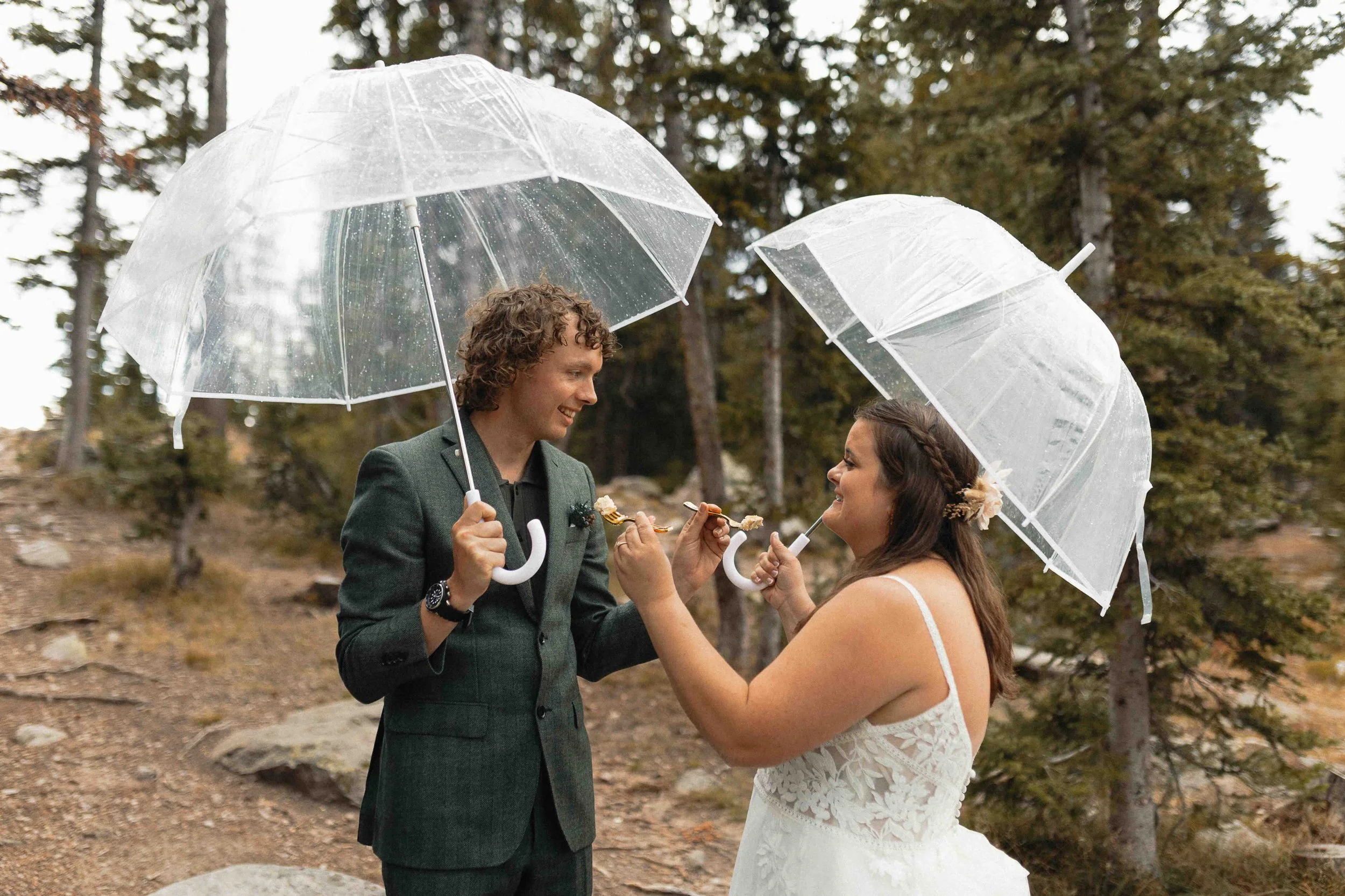 A bride and groom feeding each other cake in the forest while holding clear umbrellas.