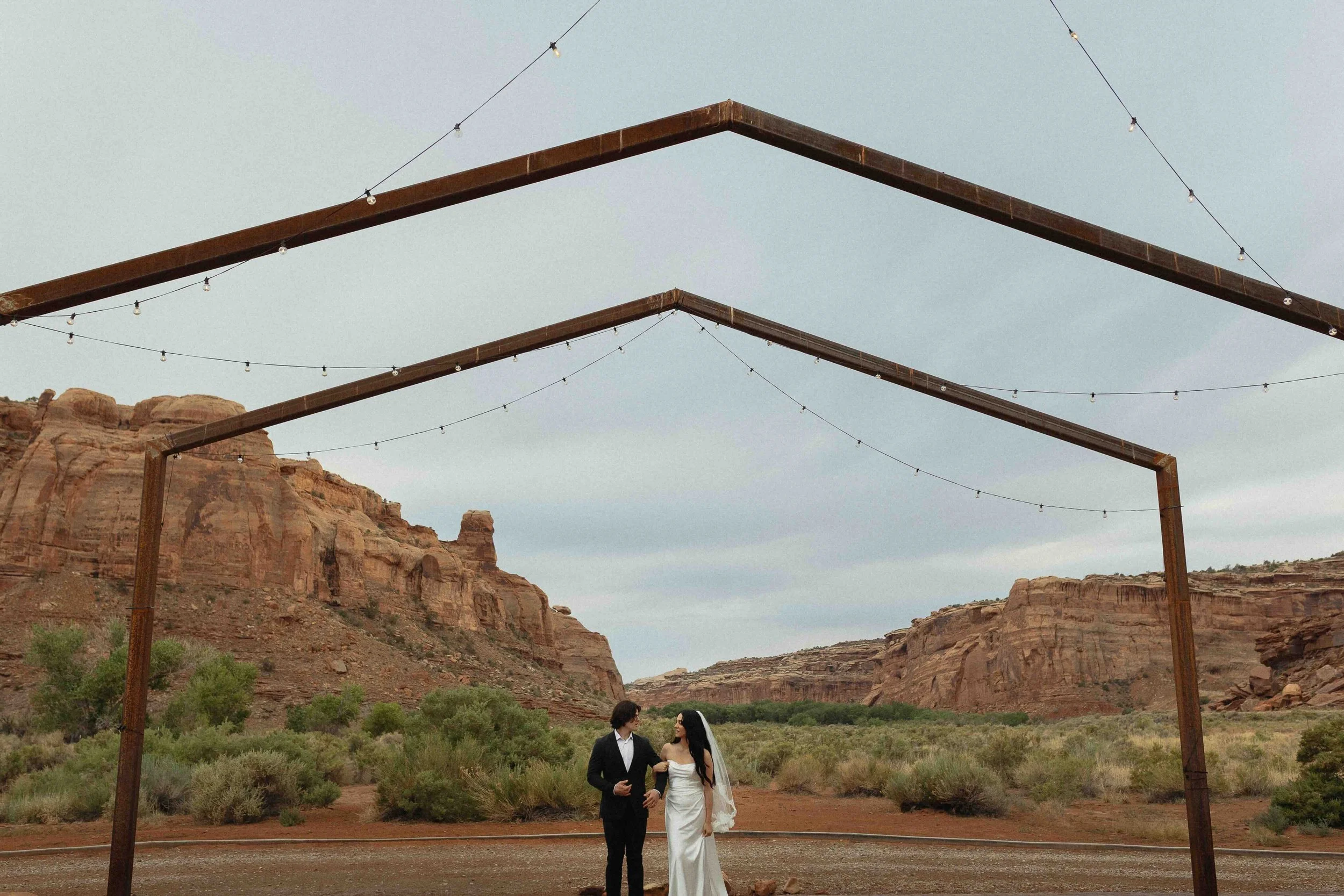 Groom escorting his bride under metal arches with desert Moab landscape in the background.