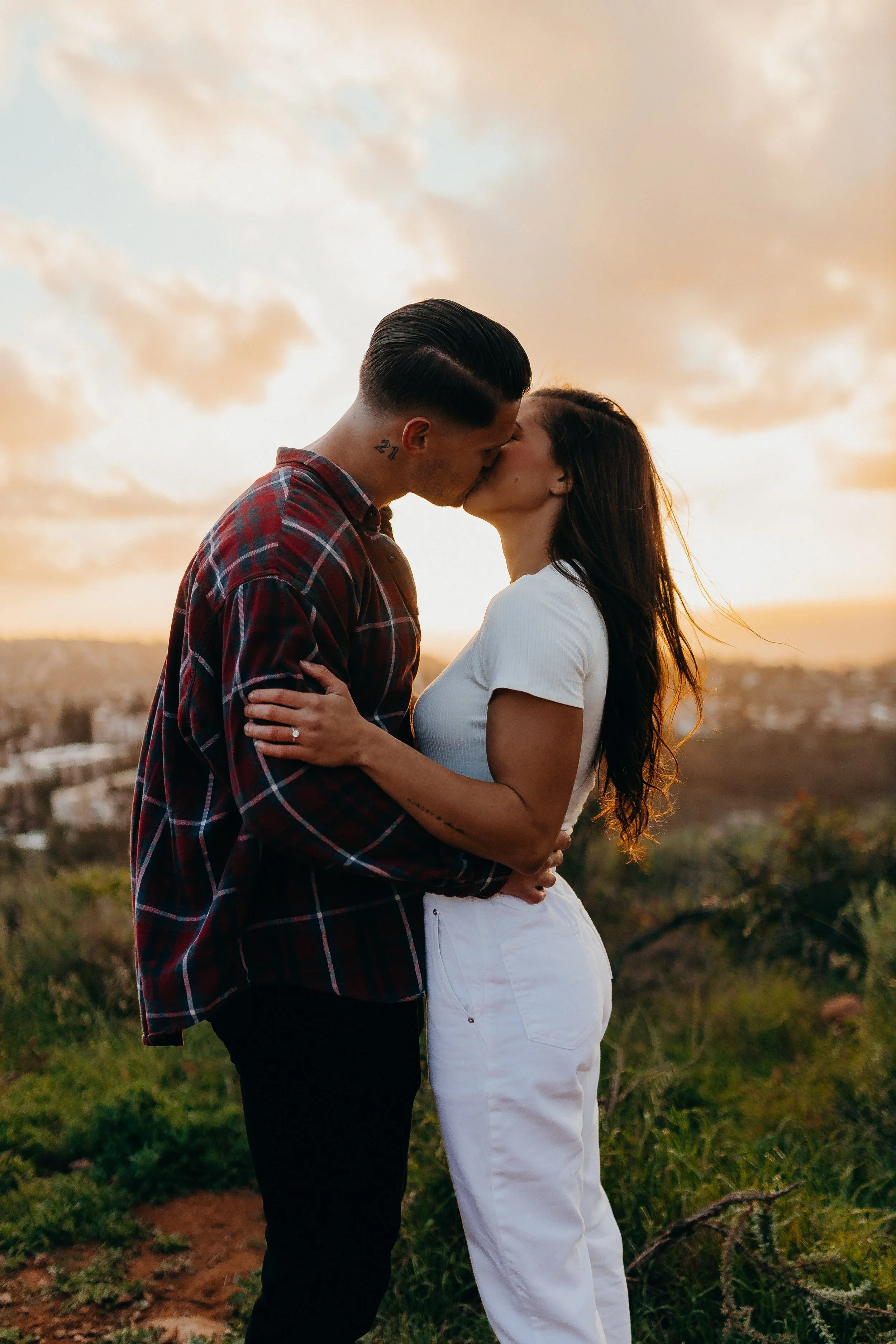 A man and woman sharing a kiss at sunset outdoors, with a cityscape in the background.