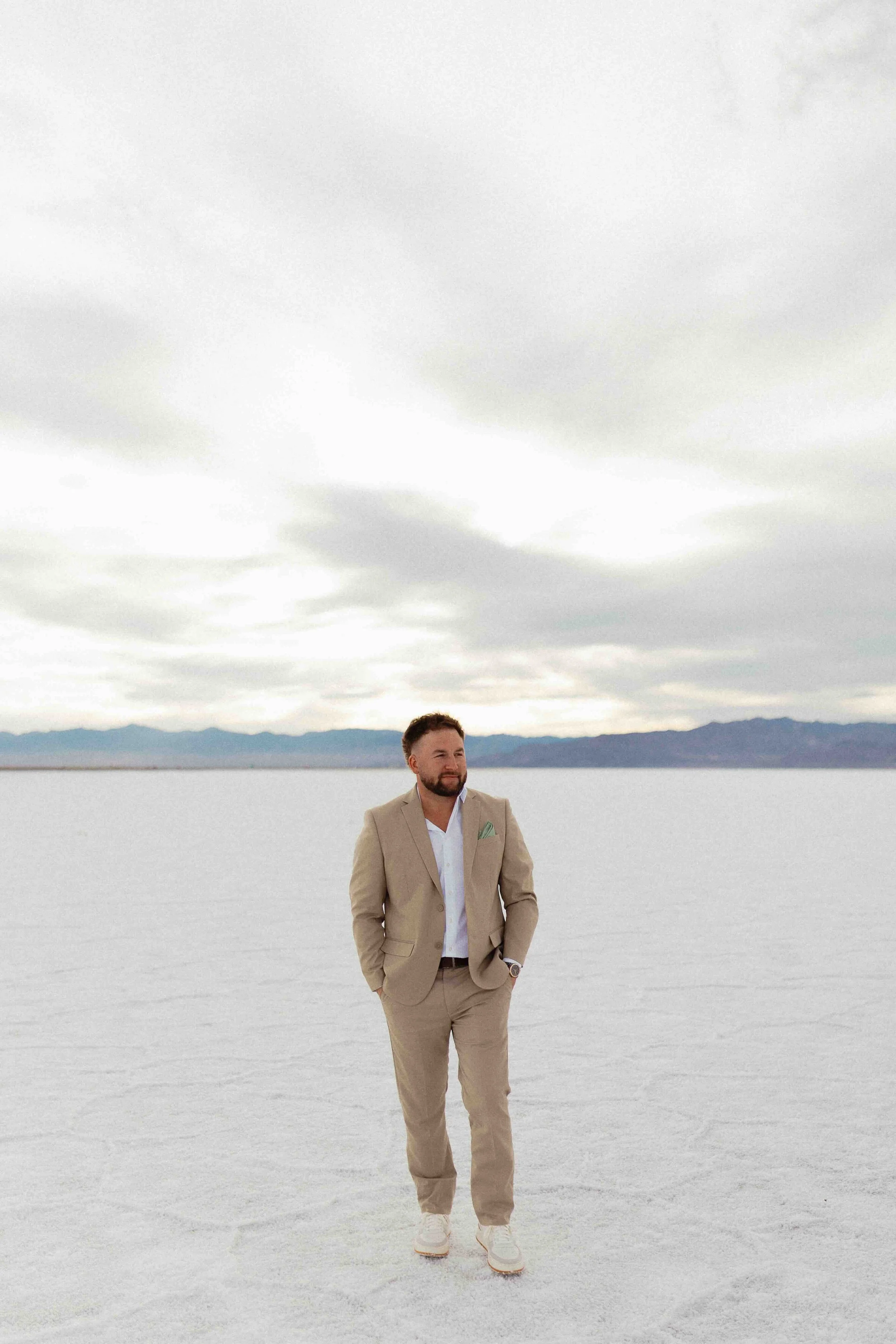 Groom in tan suit standing of the Bonneville salt flats of utah with mountains in the background.