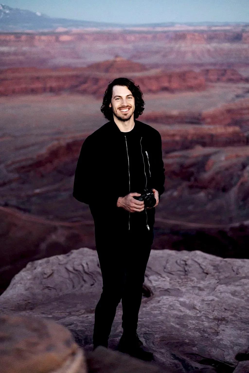 A man with dark curly hair and a beard, smiling, standing on a rock in the Grand Canyon, holding a camera, with the canyon's layered red and purple rock formations in the background.