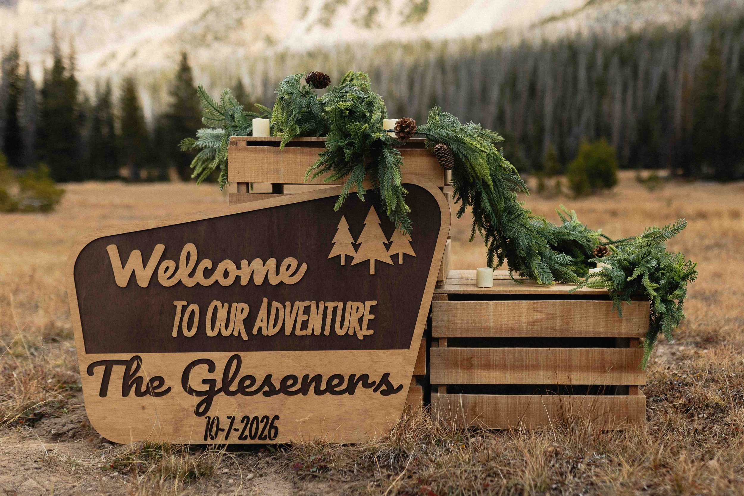 A " Welcome to our adventure" sign with pine needles on crates in front of a mountain.