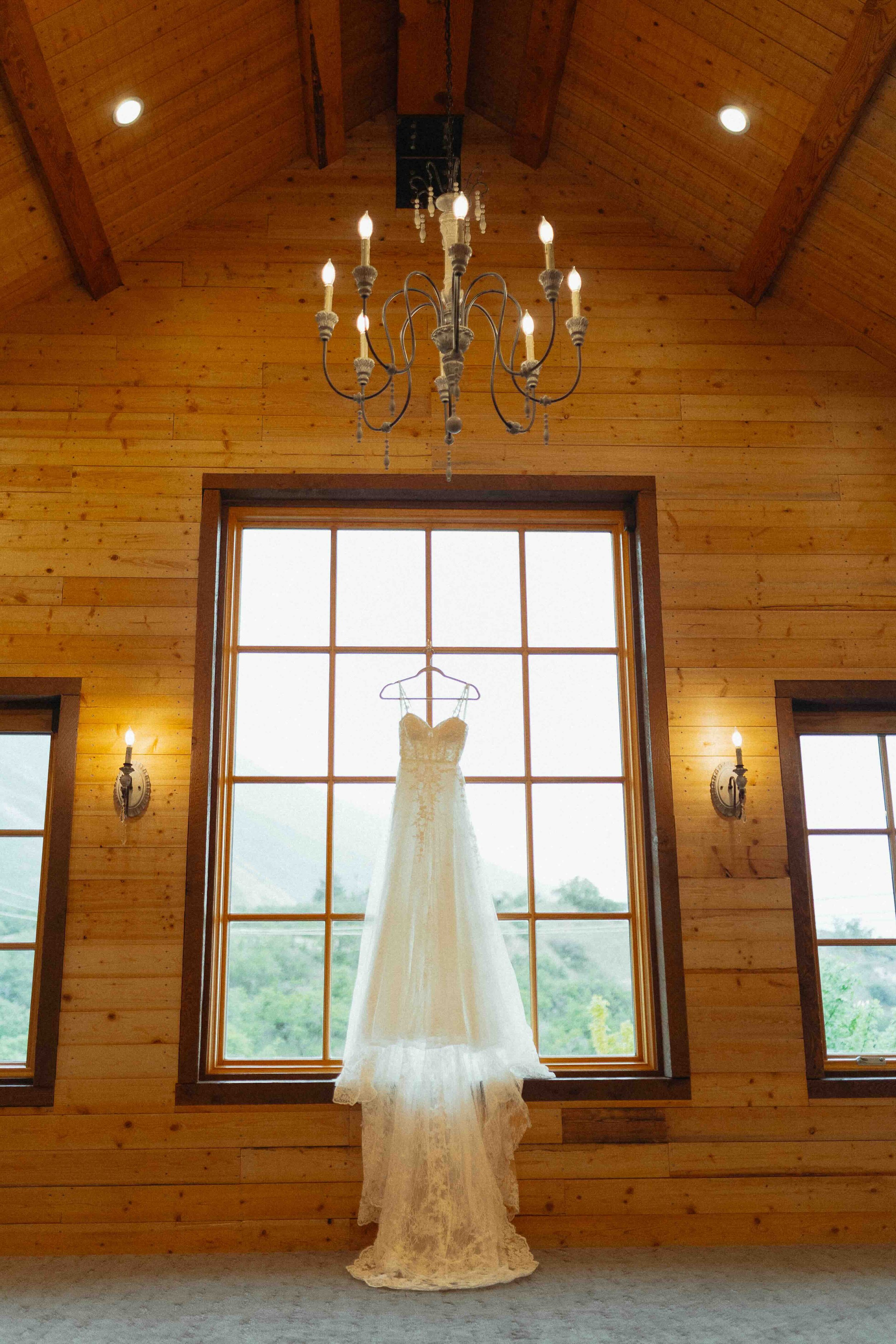 Wedding dress hanging up in a big large barn window with a chandelier above it.