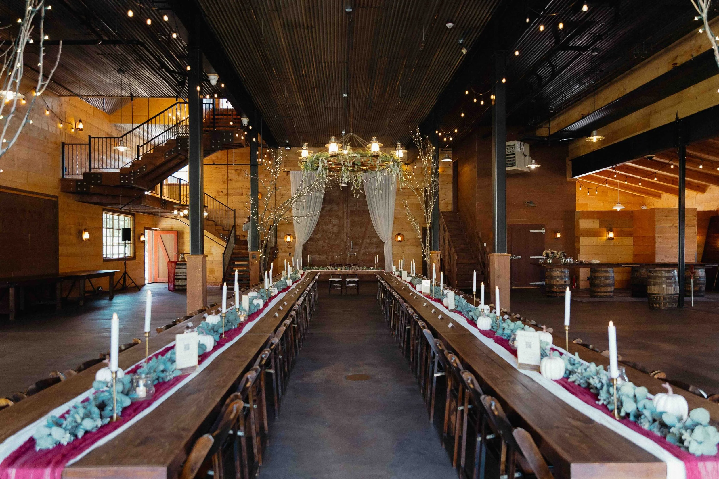 A rustic barn full of decorated tables of red table runners and white pumpkins and candlesticks in the center..