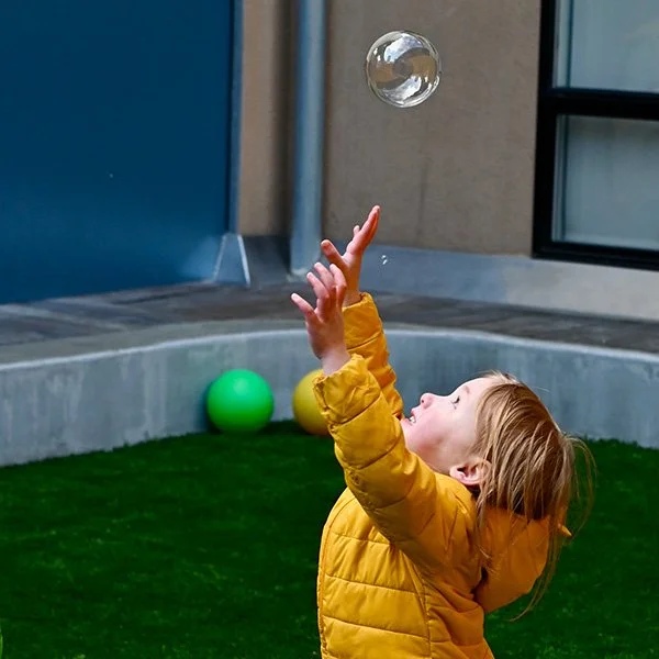 A toddler in a yellow coat plays with a bubble